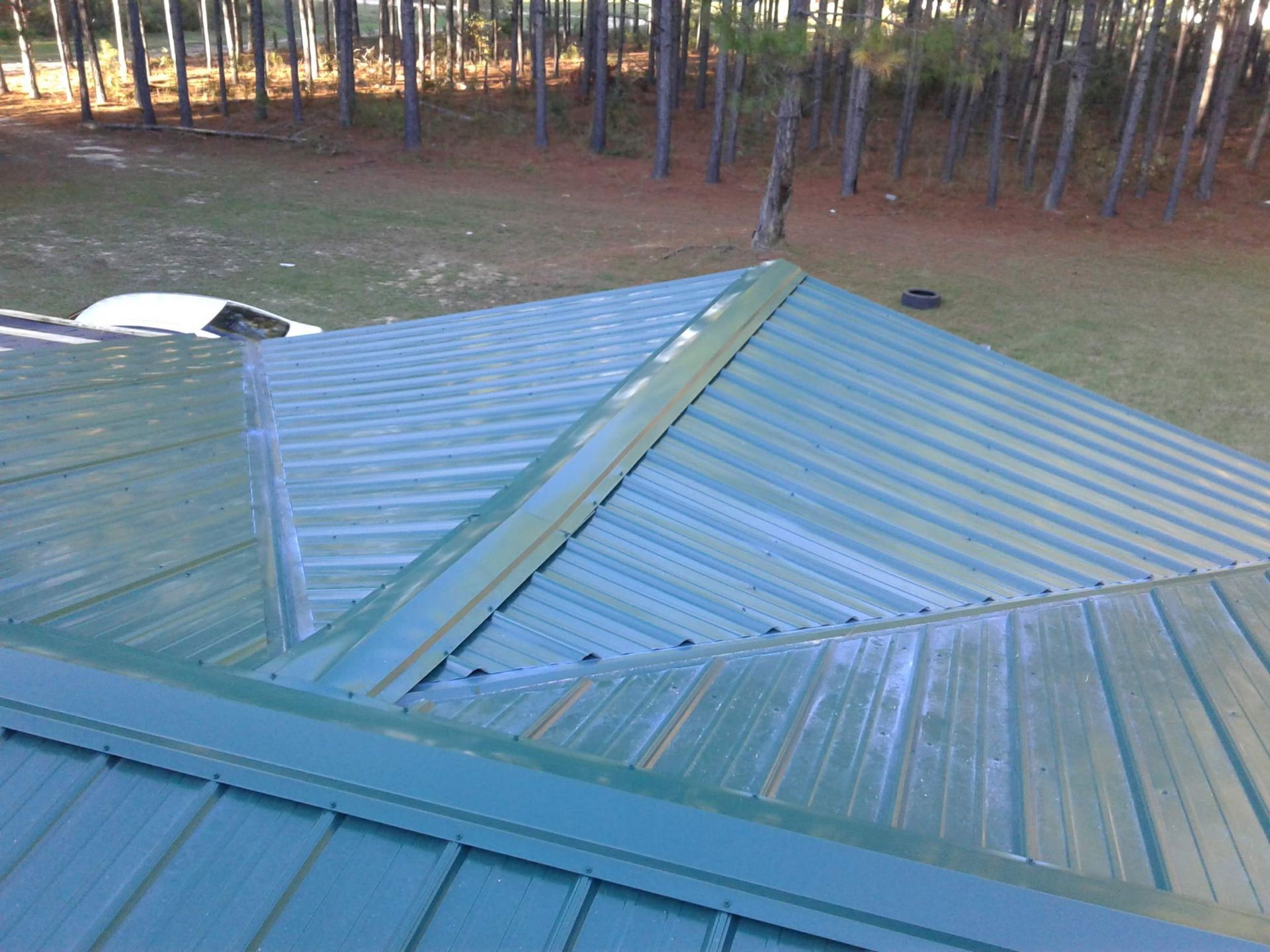 A high-angle view of a green metal roof with multiple sloped sections and valleys, set against a background of trees.