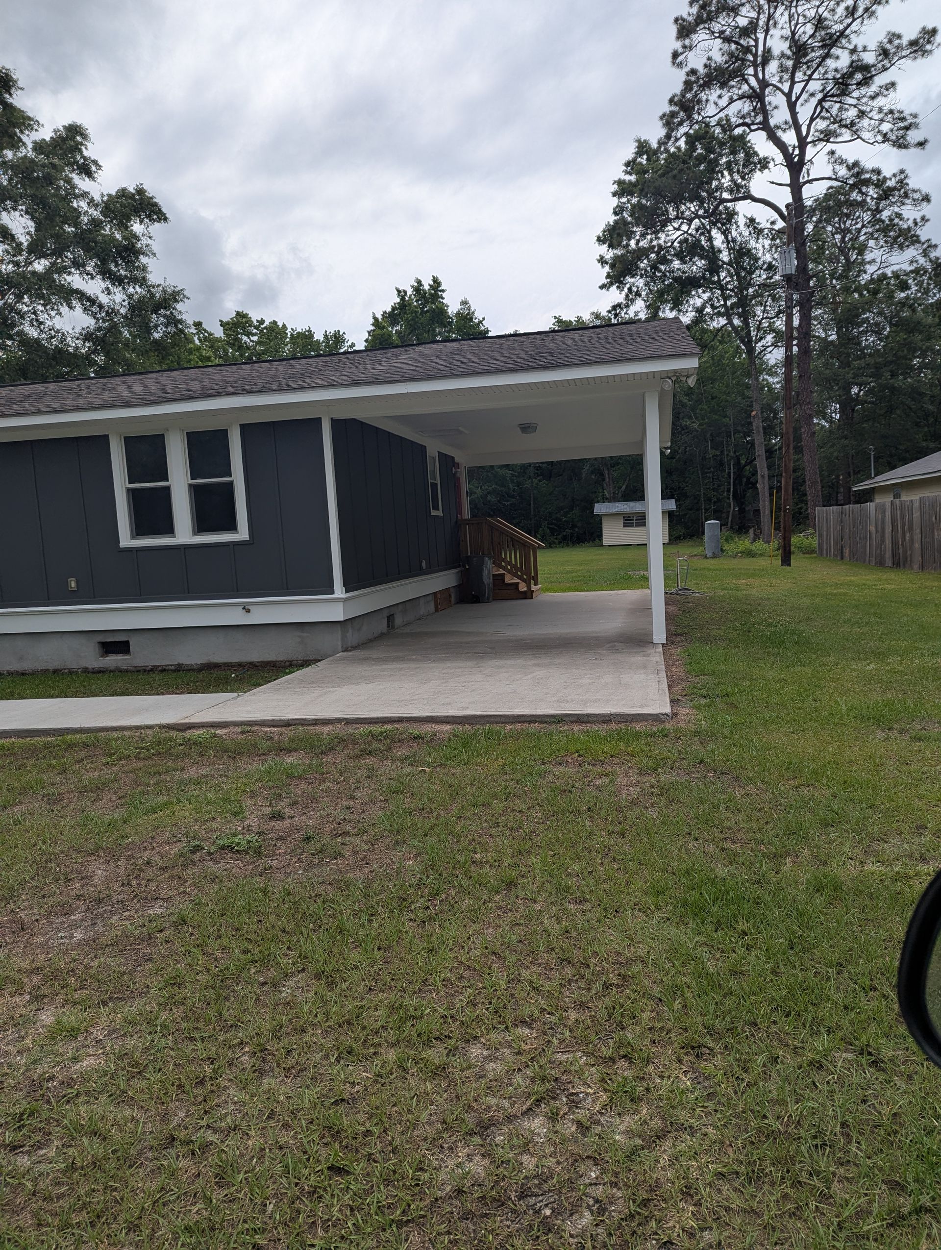 A dark gray single-story house with a white trim and a concrete carport on a grassy lot under a cloudy sky.