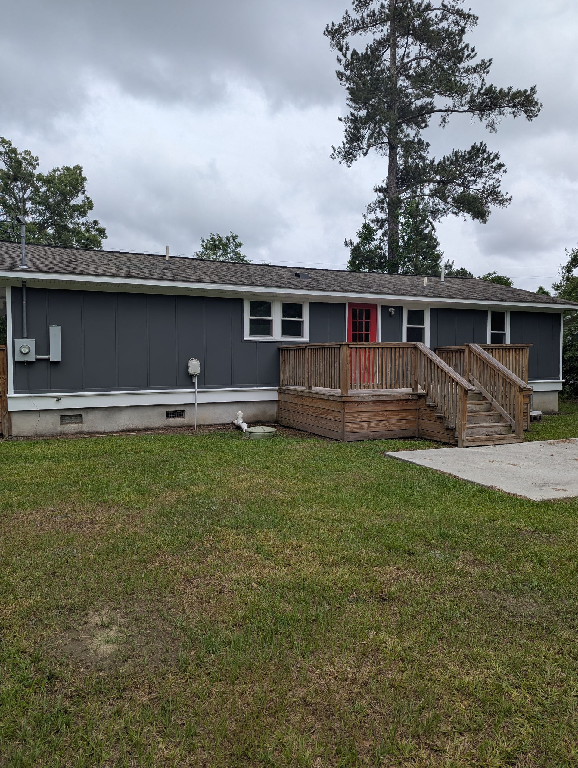 A dark gray house with a red door and a wooden deck sits on a grassy lawn under a cloudy sky.