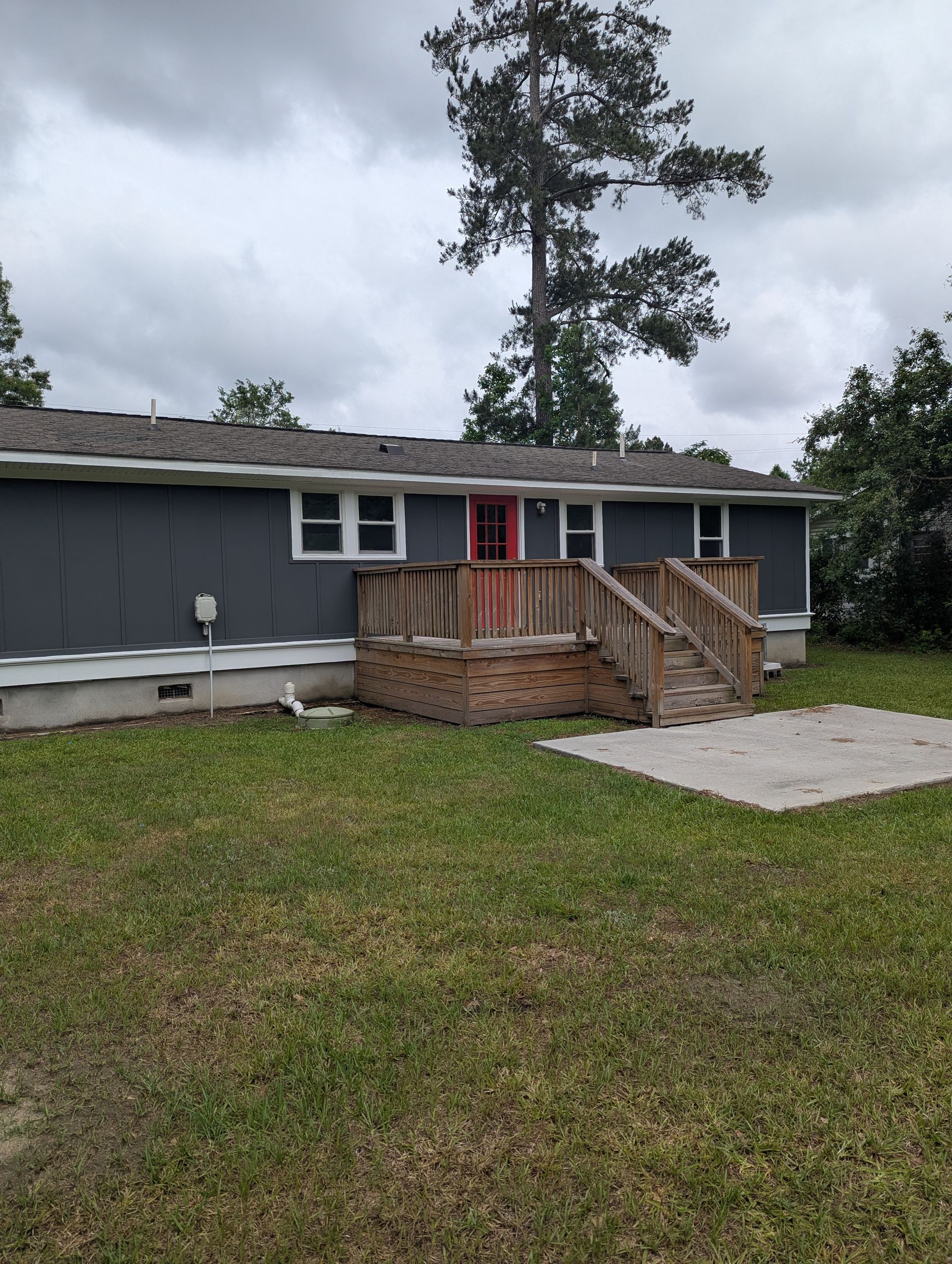 A single-story gray house with a wooden deck and a red door sits on a grassy lot under a cloudy sky.