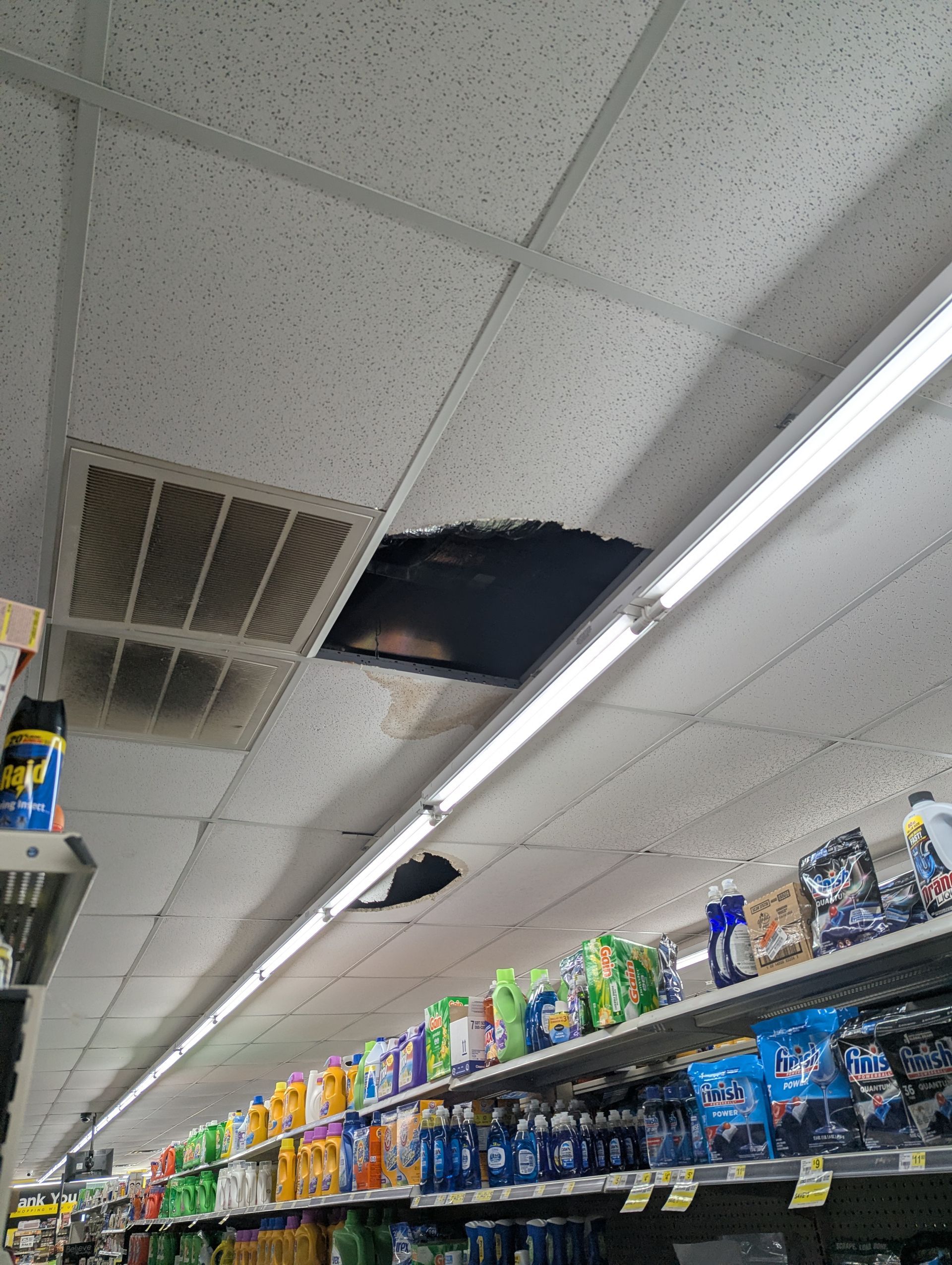 Damaged ceiling tiles with visible black holes and soot inside a retail store aisle stocked with cleaning supplies.