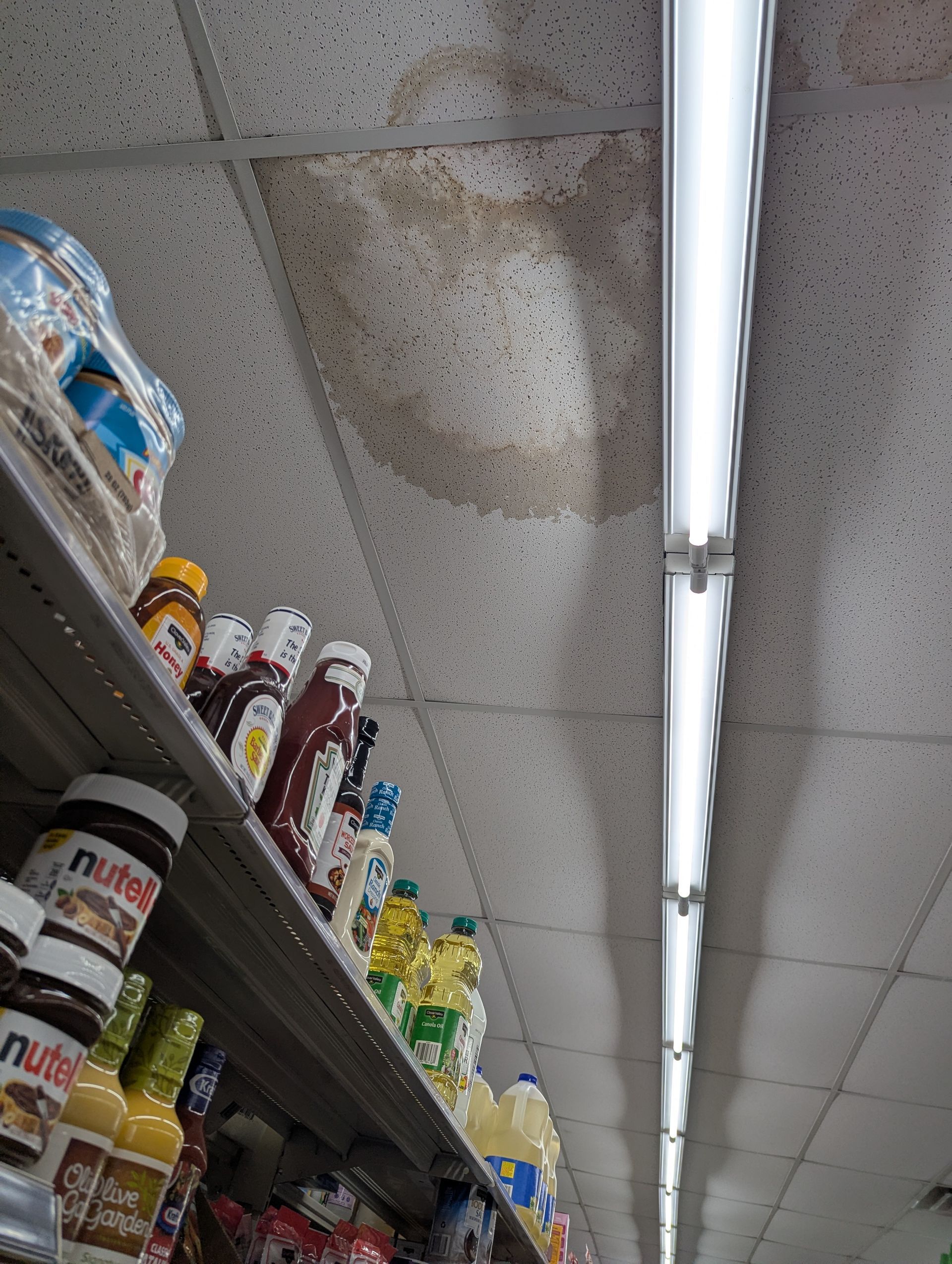 Large water stain on a store's ceiling tiles above a shelf stocked with food products and a long light fixture.