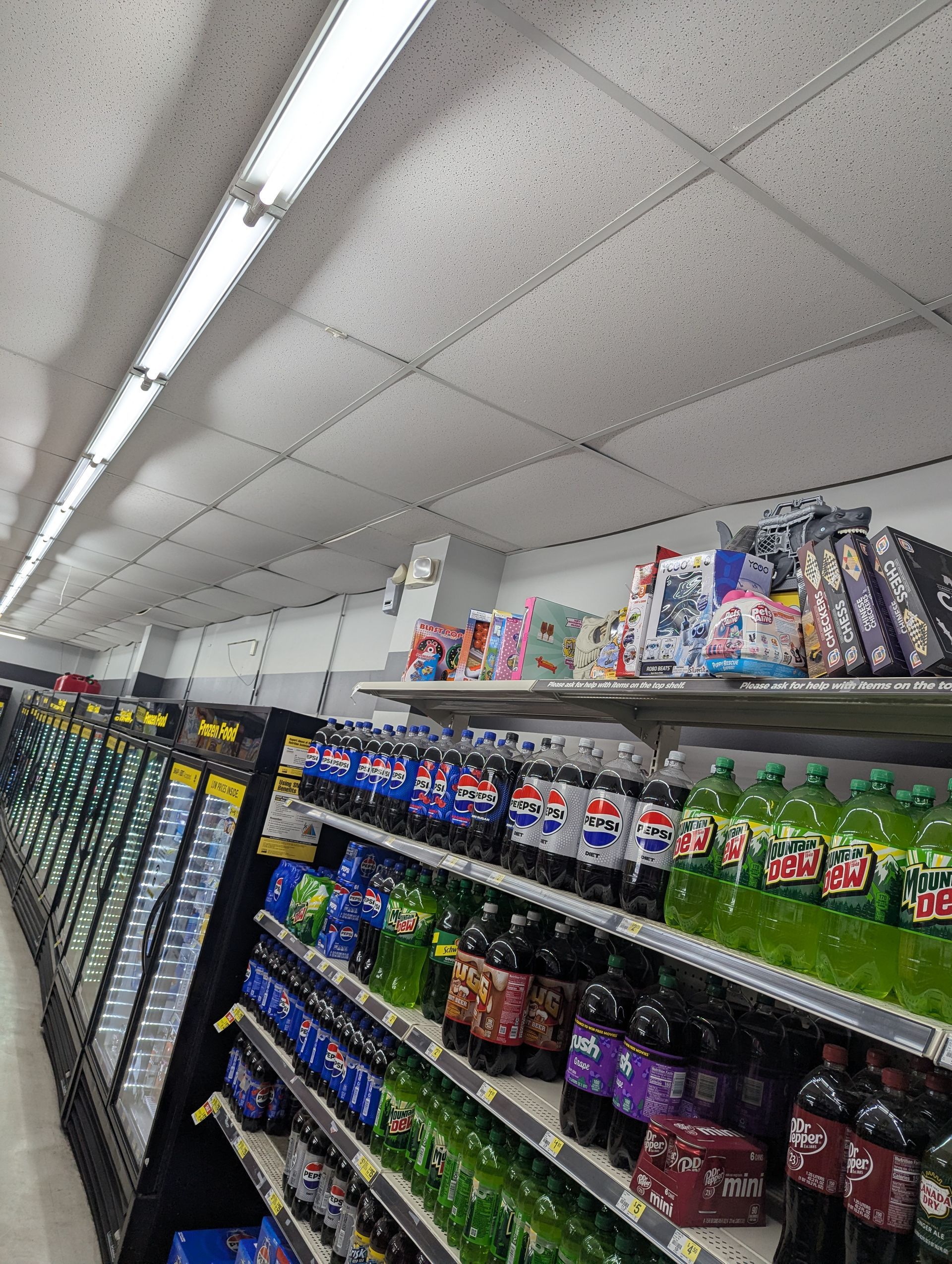 Rows of assorted plastic soda bottles on retail shelves inside a store, with boxed products displayed on the top shelf.