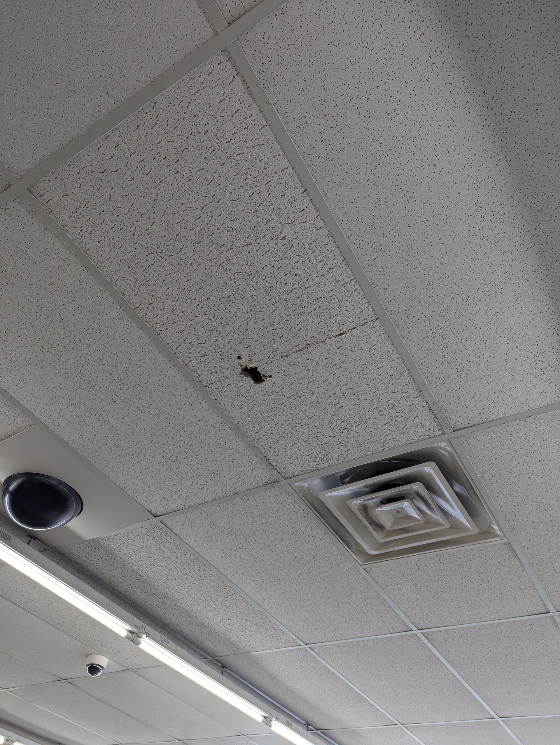 A drop ceiling panel features a small hole, positioned near a square ventilation grate and an indoor security camera.