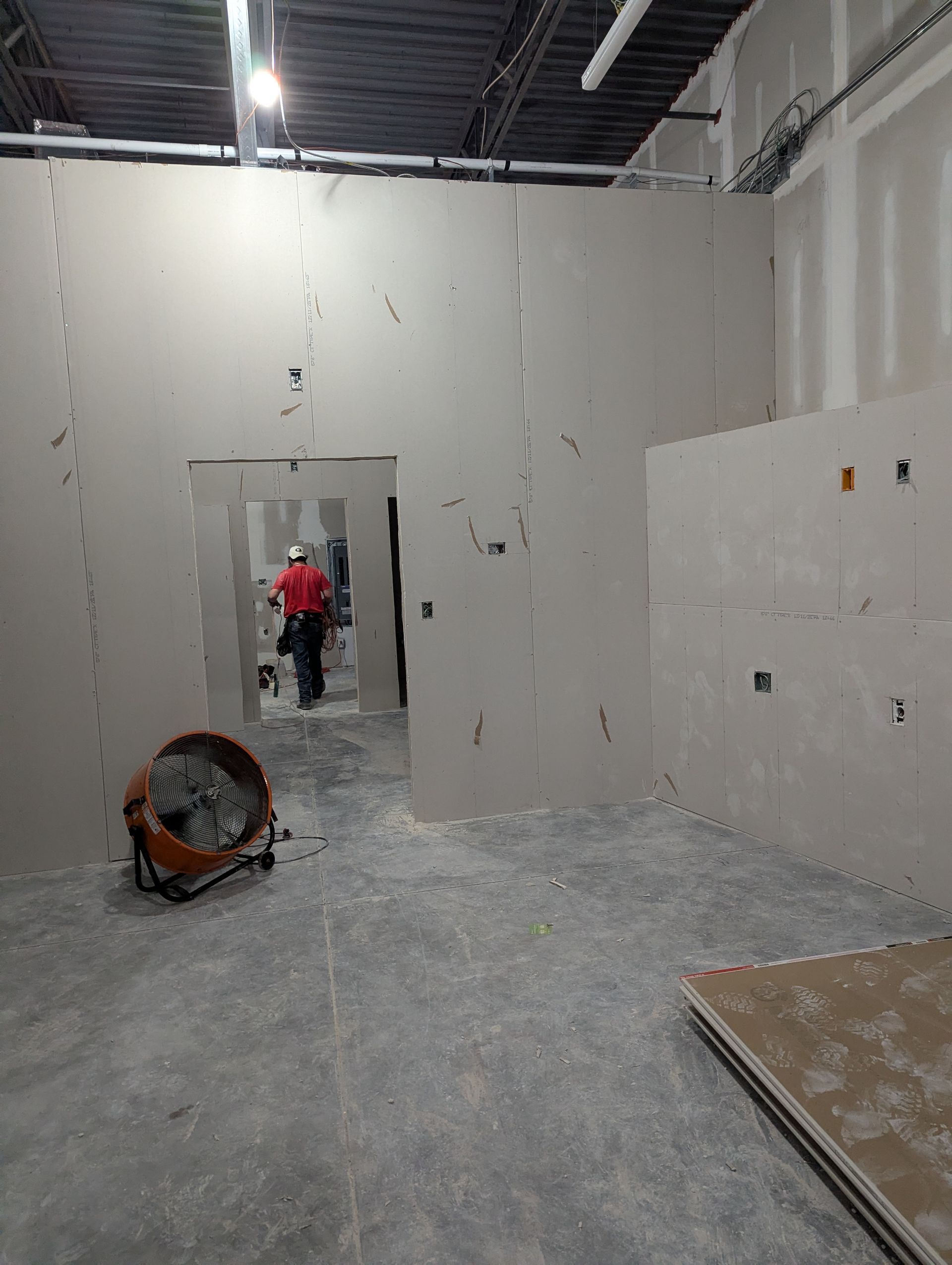 An interior construction site with drywall-finished walls, a doorway leading to a back room, and a person walking through.