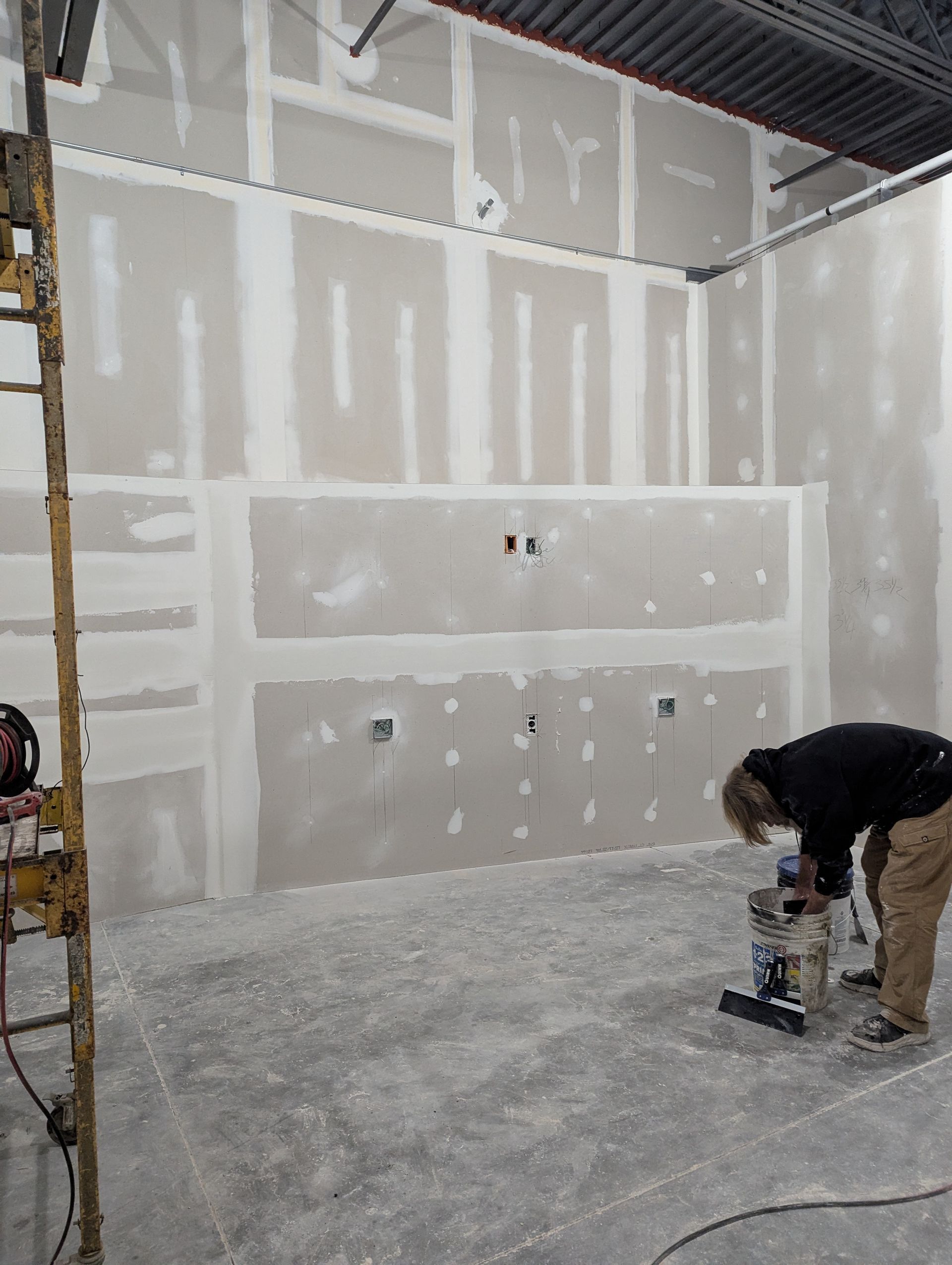 A person using a long-handled tool to apply compound to a taped drywall seam in an unfinished building.