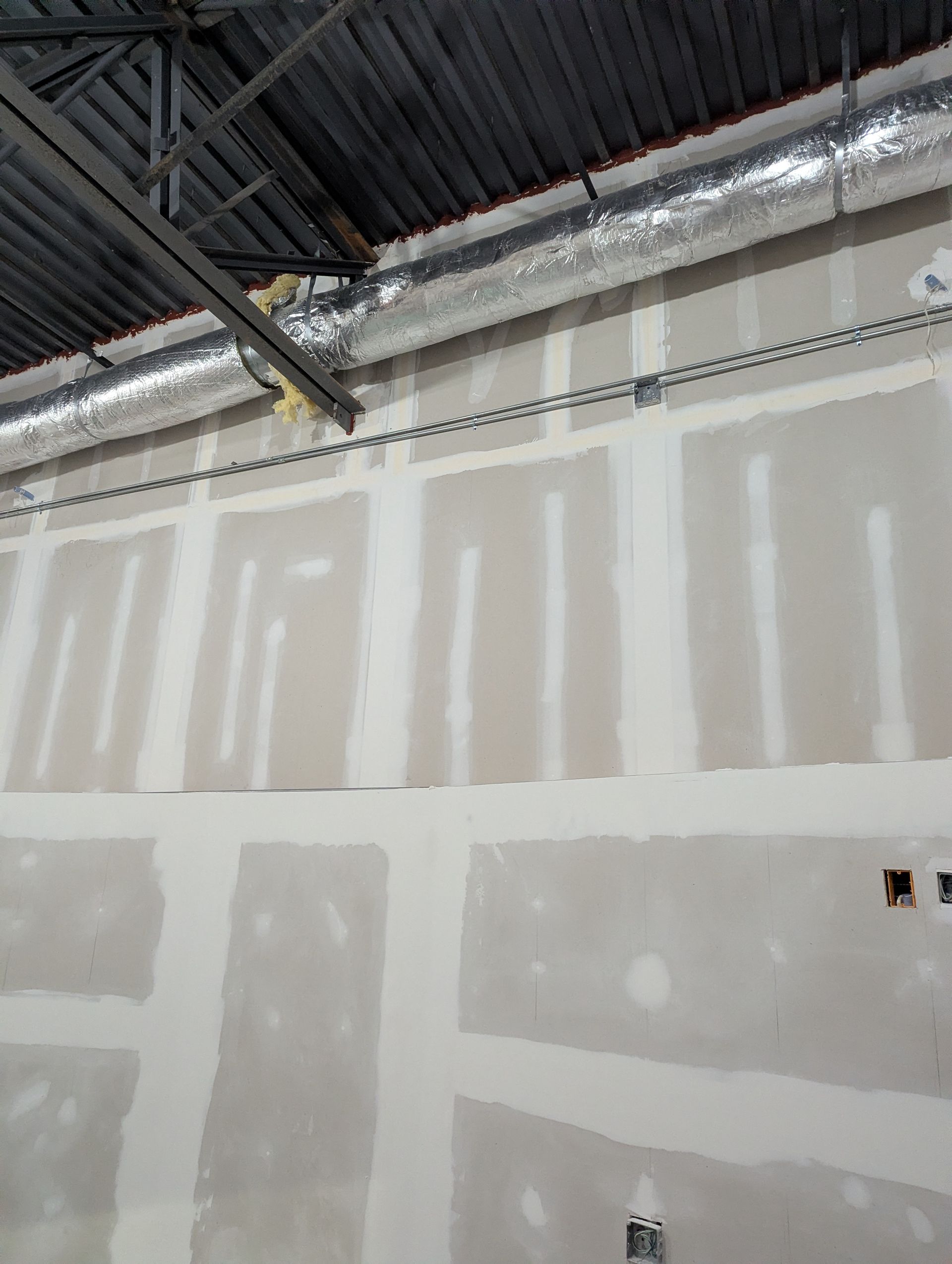 An interior wall under construction with unfinished drywall, taped seams, exposed metal ductwork, and ceiling beams.