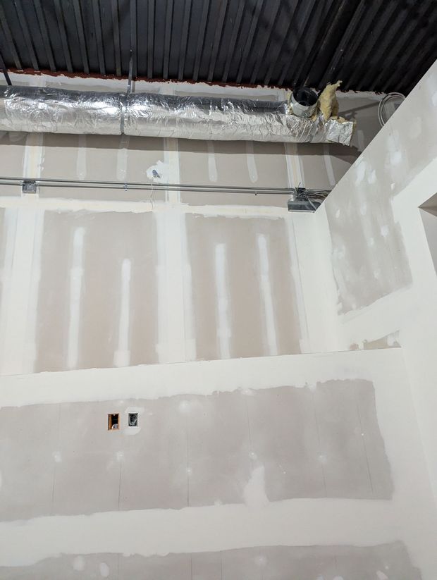 Unfinished interior wall with taped drywall, exposed metal ductwork, and electrical wiring on a corrugated ceiling.