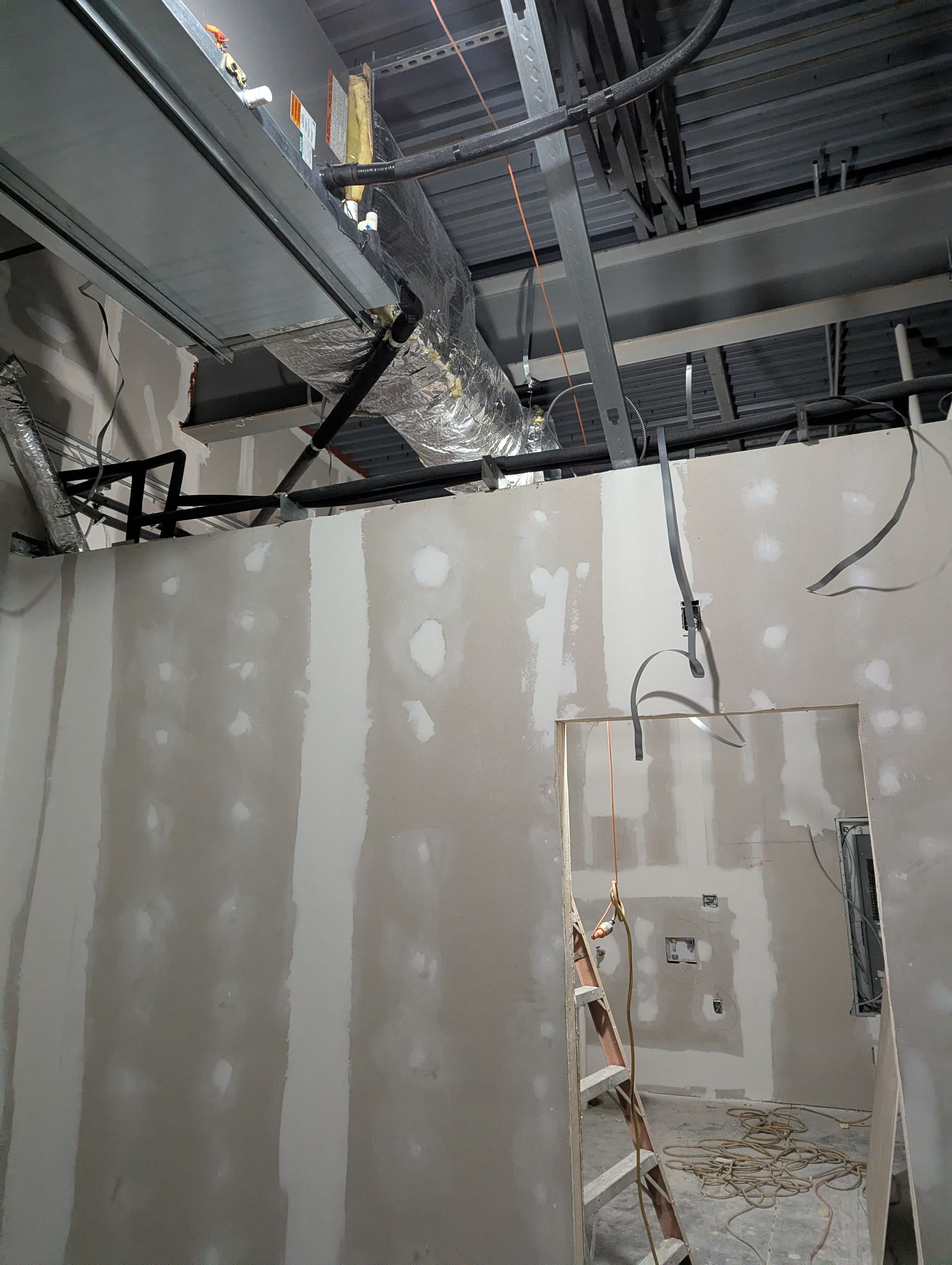 Interior construction site with unfinished drywall partitions, exposed ceiling pipes, and a ladder in the doorway.