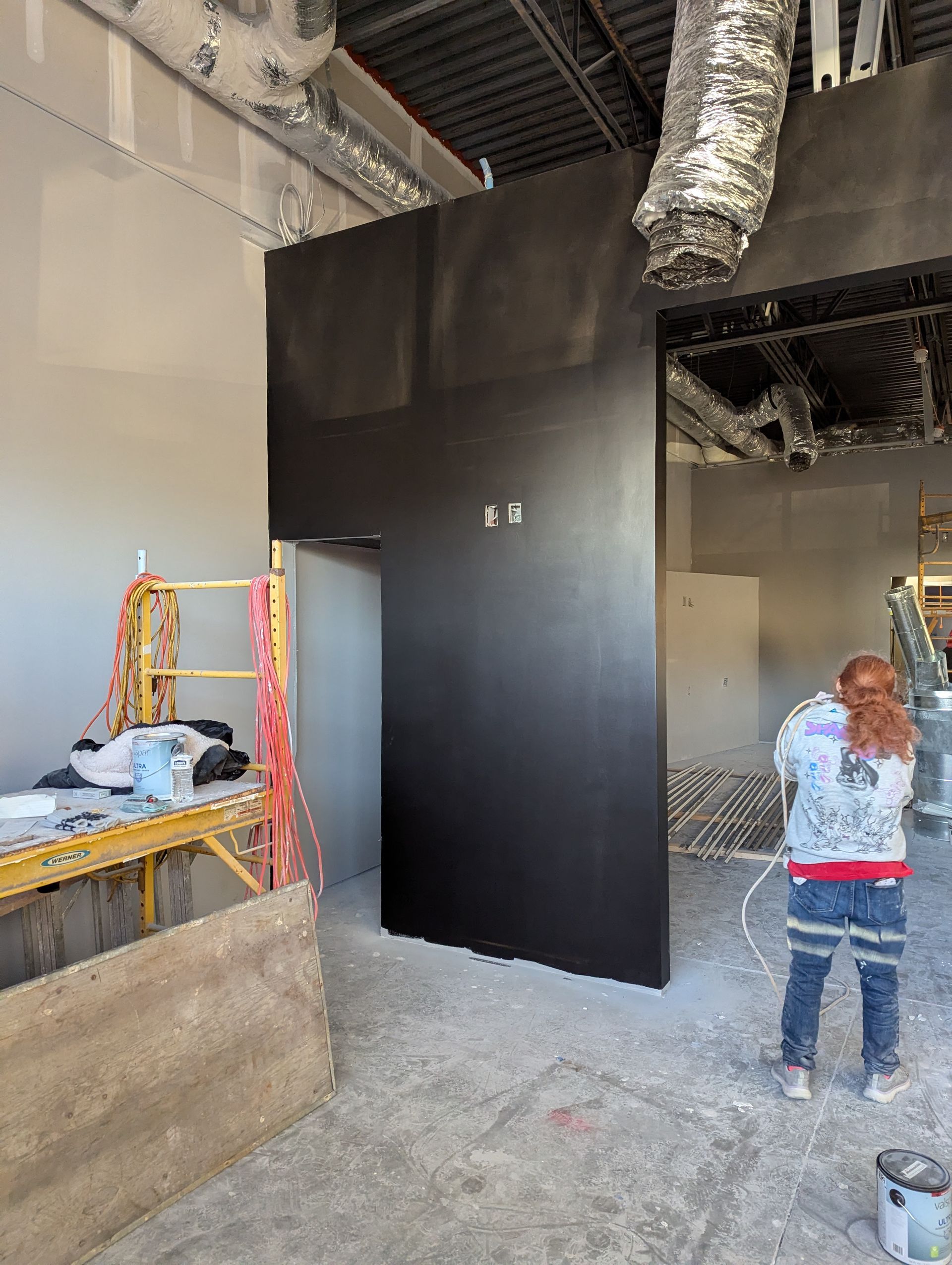 A person in work clothes stands in an unfinished room next to a wall painted matte black, with scaffolding to the left.