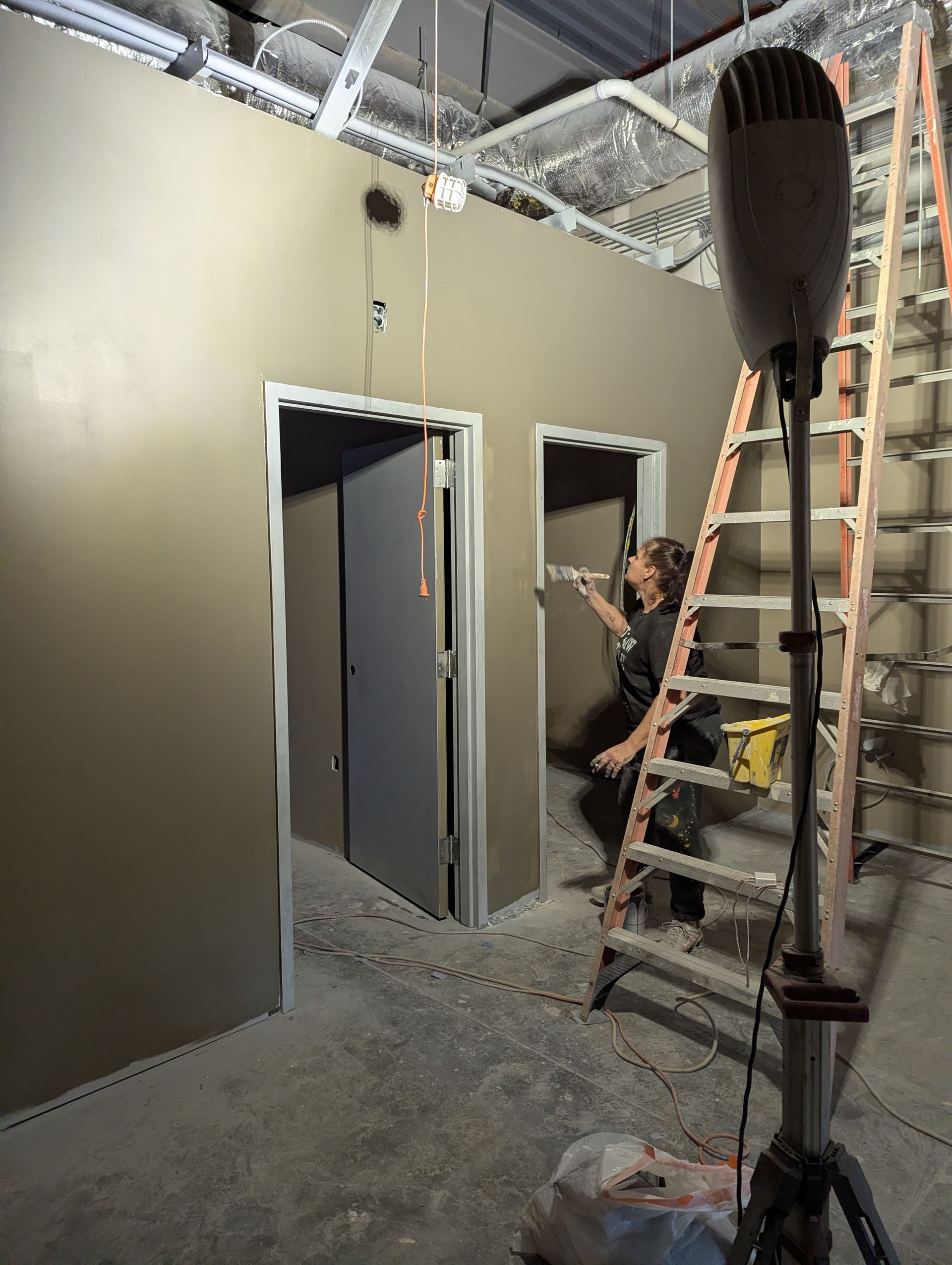 A person paints a doorway frame in a room under construction, with a tall work light and ladder nearby.