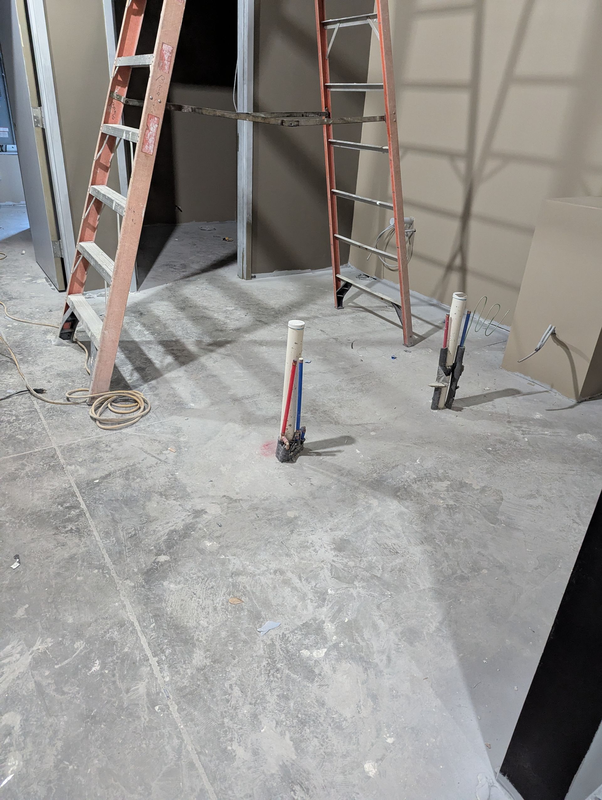 Construction area with two step ladders and two plumbing pipes protruding from a concrete floor.