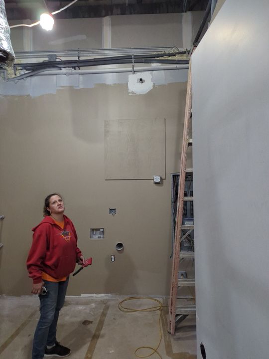 A person in a red sweatshirt holding a tool stands in an unfinished room with drywall, an exposed ceiling, and a ladder.