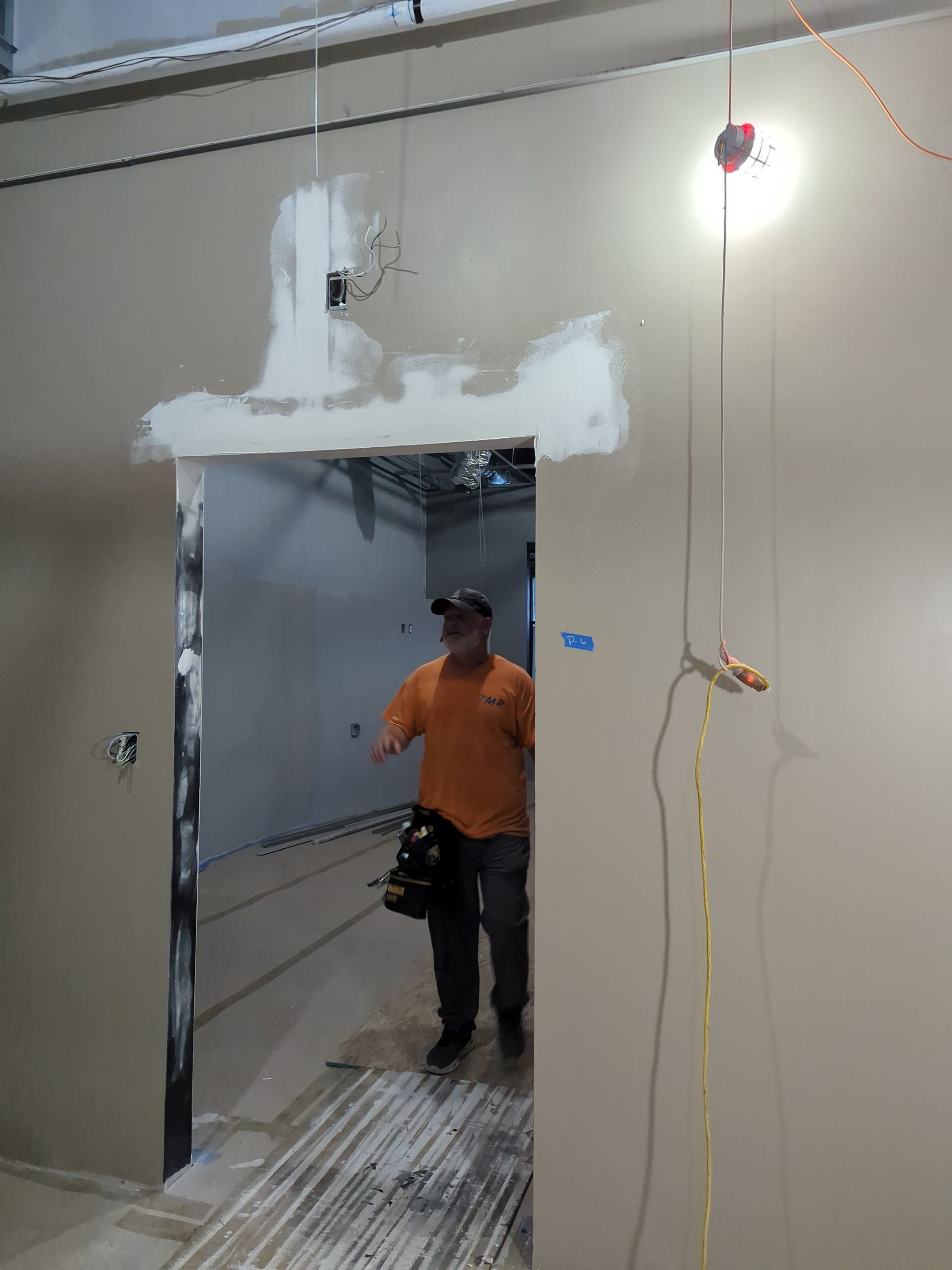A person in an orange shirt walks through an unfinished doorway in a room with tan walls and exposed construction.