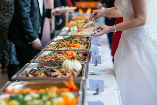 Buffet table with trays of food while a guest in a white dress serves a plate at an event