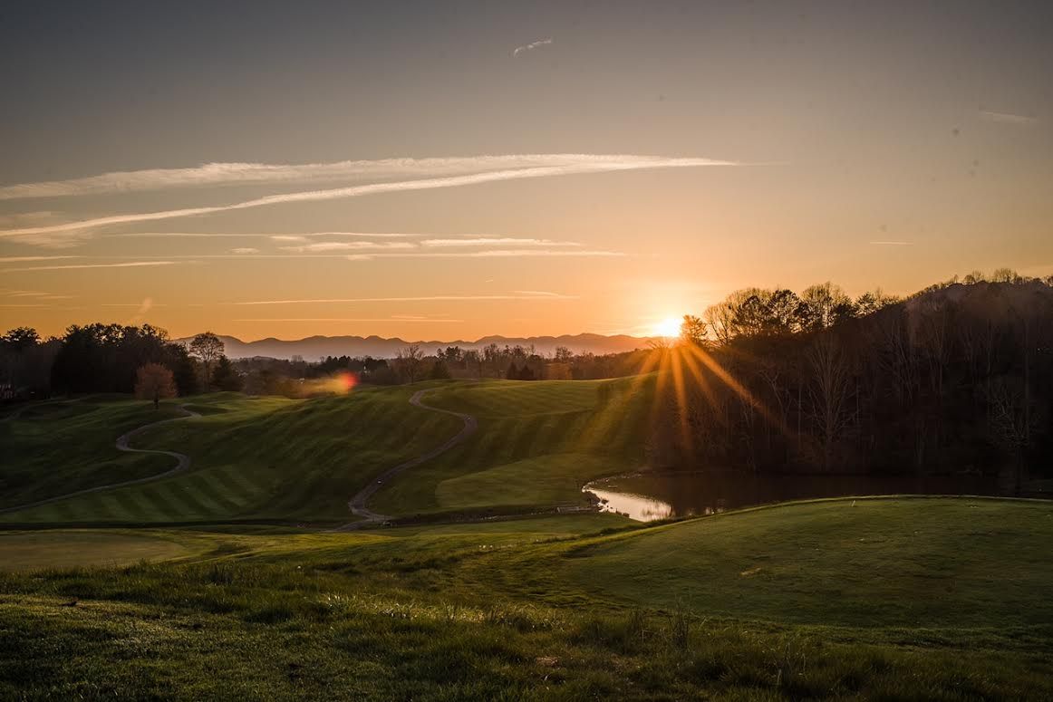 Sunset over a green golf course with a small creek and silhouetted trees.