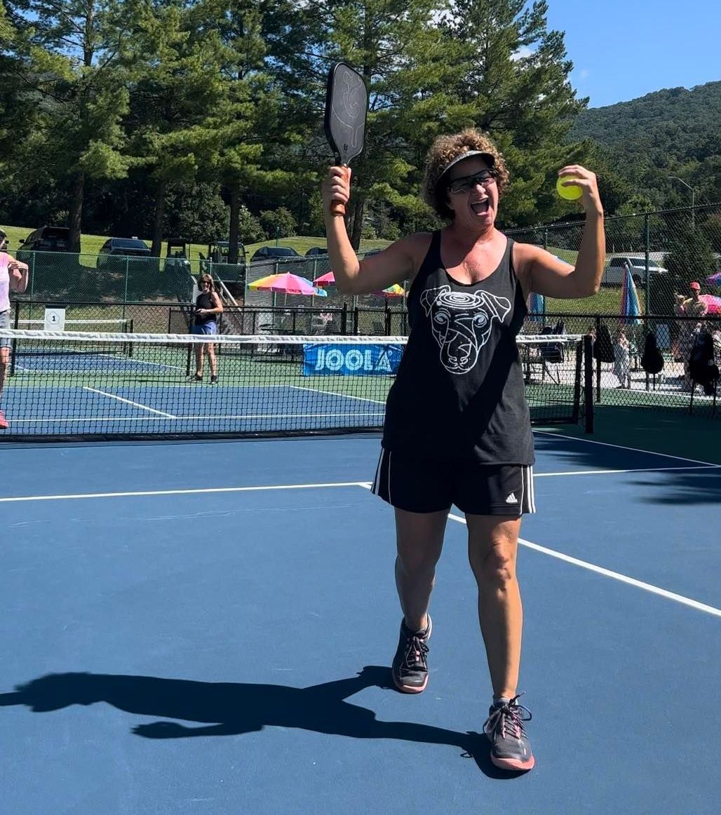 Woman celebrates pickleball victory, holding paddle and ball, on outdoor court.