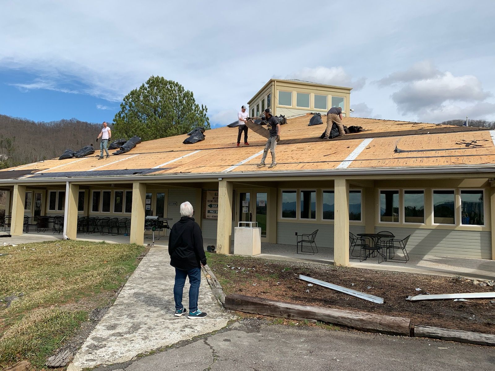 People replacing roof shingles on a building with a flat roof; person observes. Sunny day.