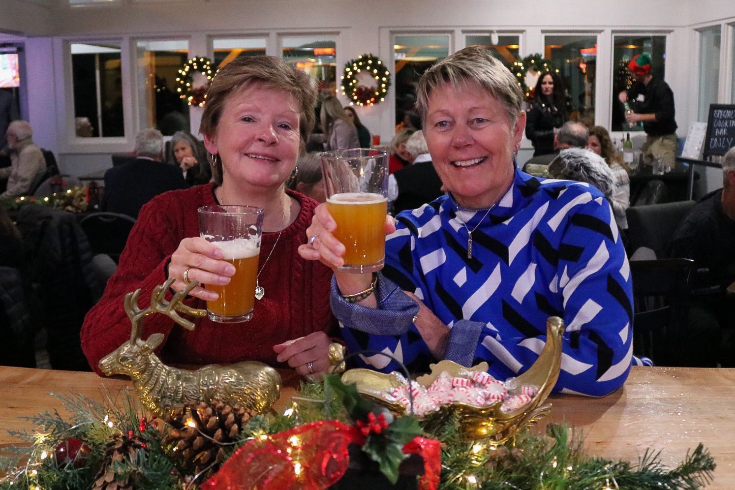 Two women toasting with beer at a bar, decorated for the holidays.