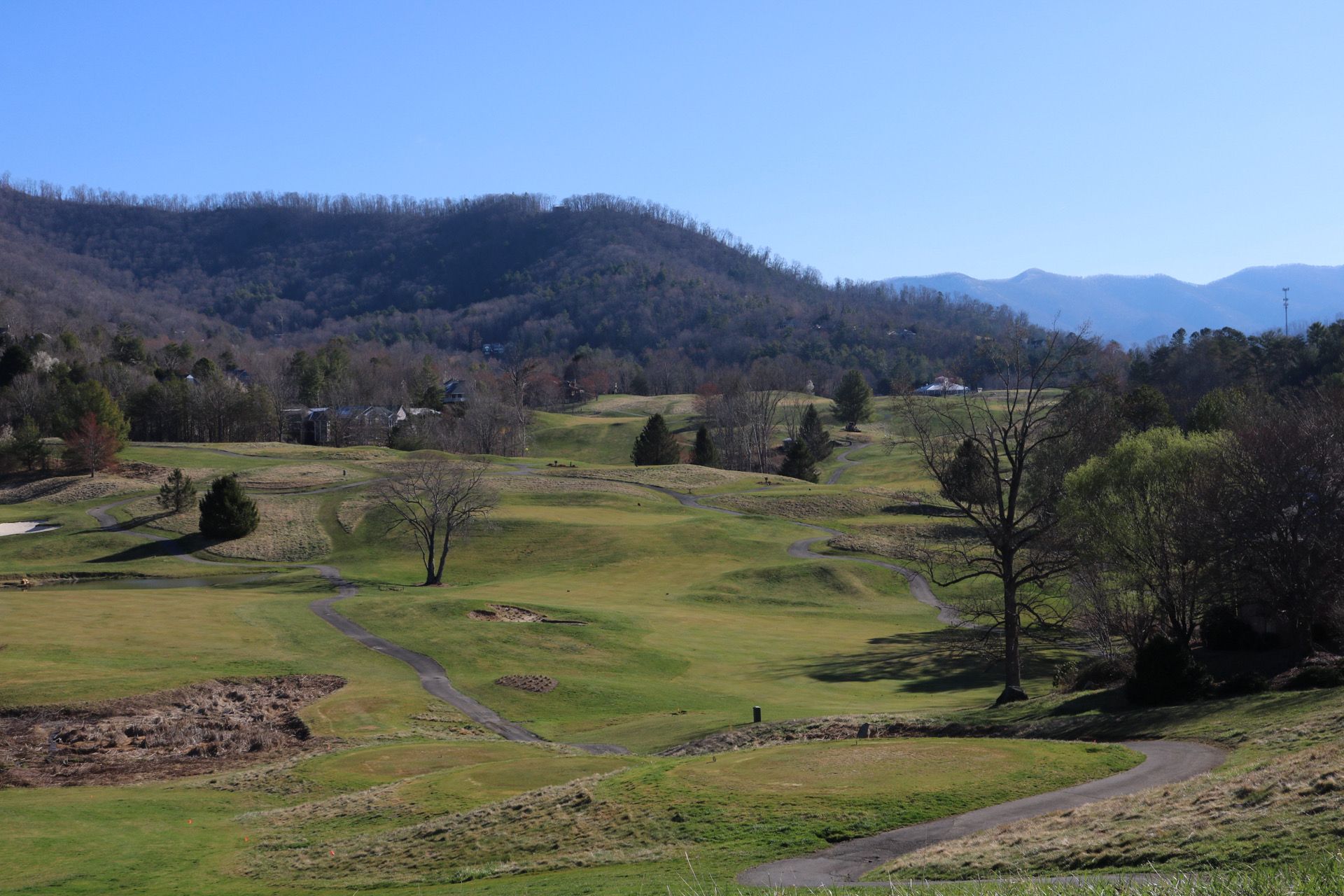 Green golf course with rolling hills, trees, and a mountain backdrop under a blue sky.