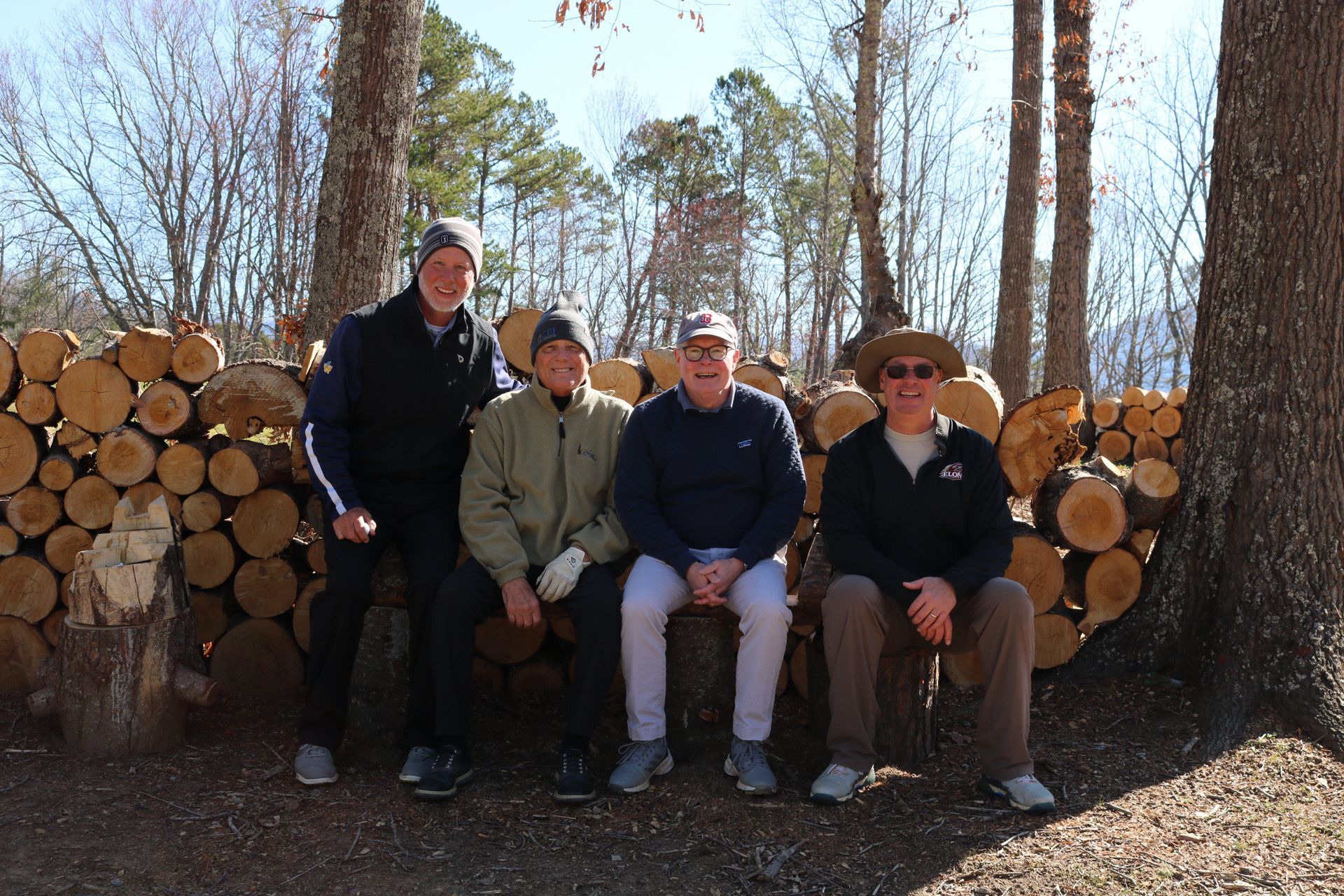 Four people sitting on a stack of logs in front of a pile of firewood, trees in background.