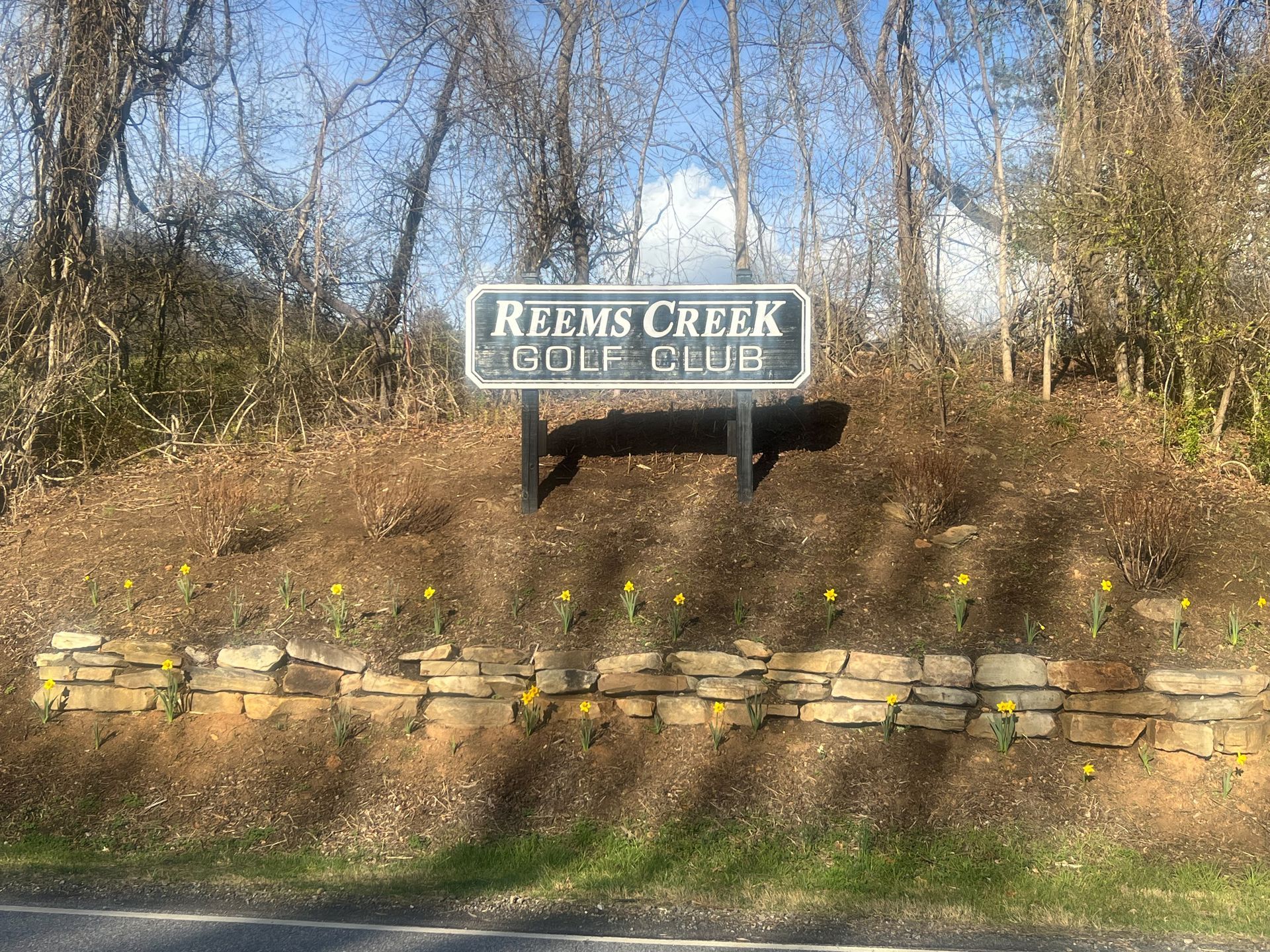 Sign for Reems Creek Golf Club, mounted on a small hill with stone wall and daffodils, trees in background.