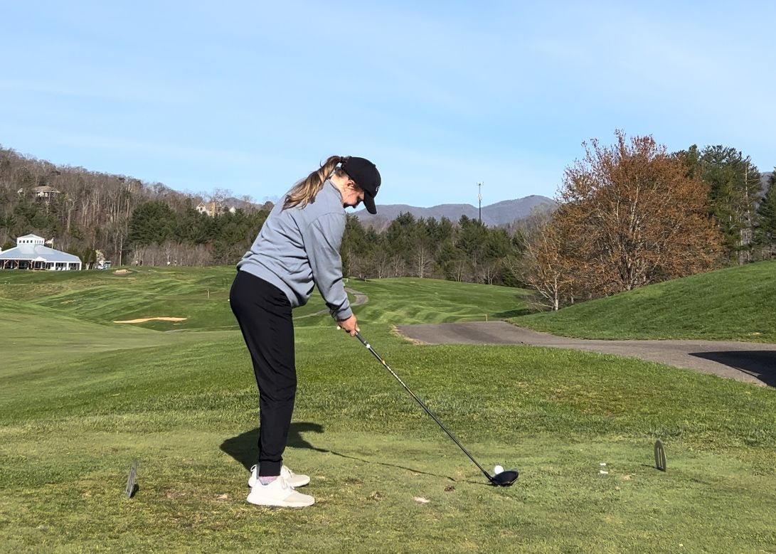 Person teeing off on a golf course, mountains in the background, sunny day.