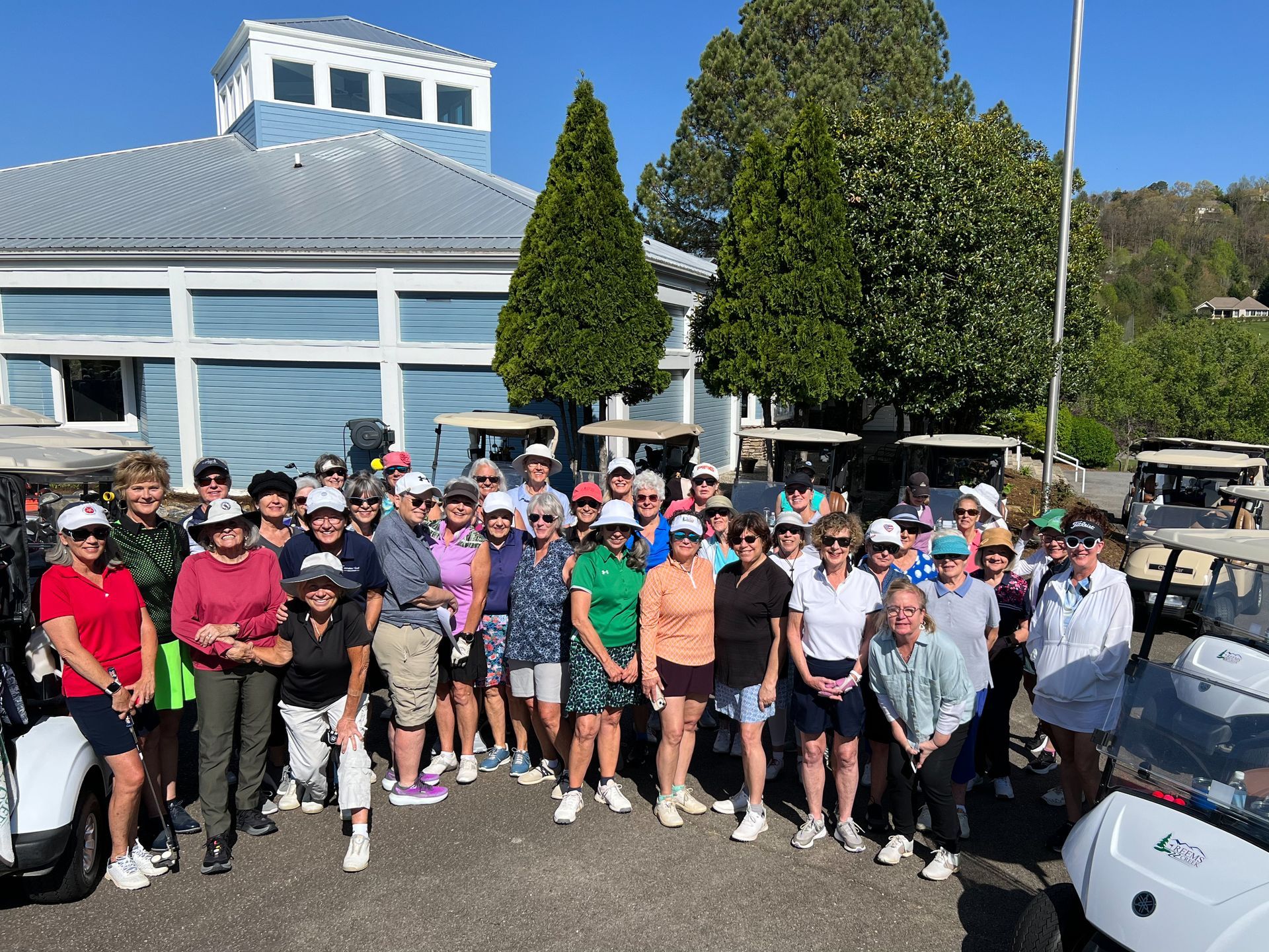 Group of golfers in front of a blue building, posed for a photo. Golf carts present.