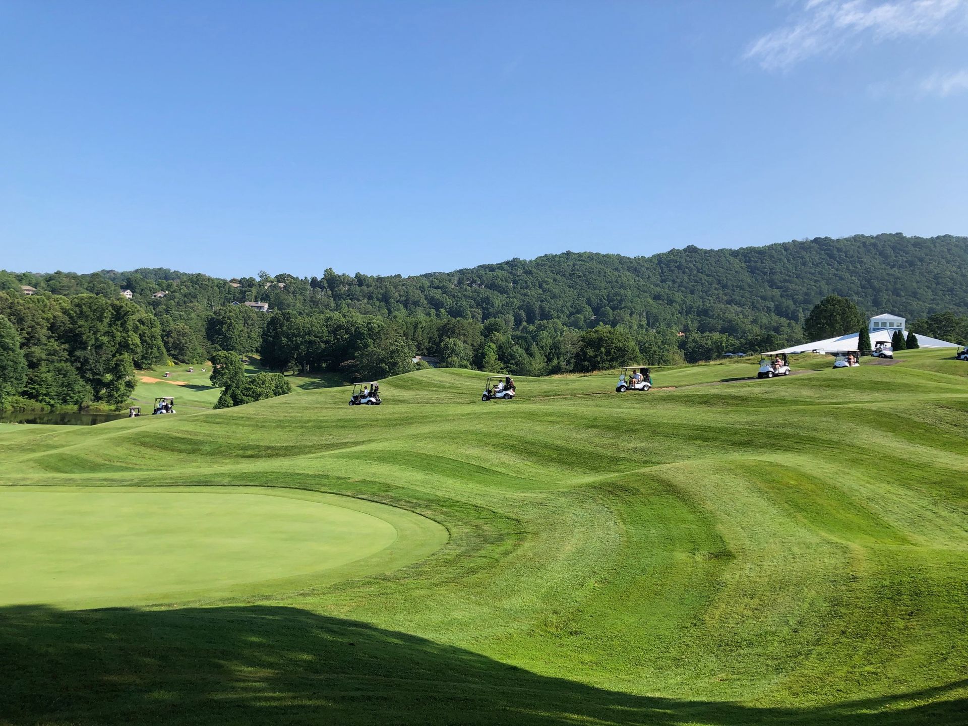 Golf course with rolling green hills, golf carts, and a tree-covered mountain under a blue sky.
