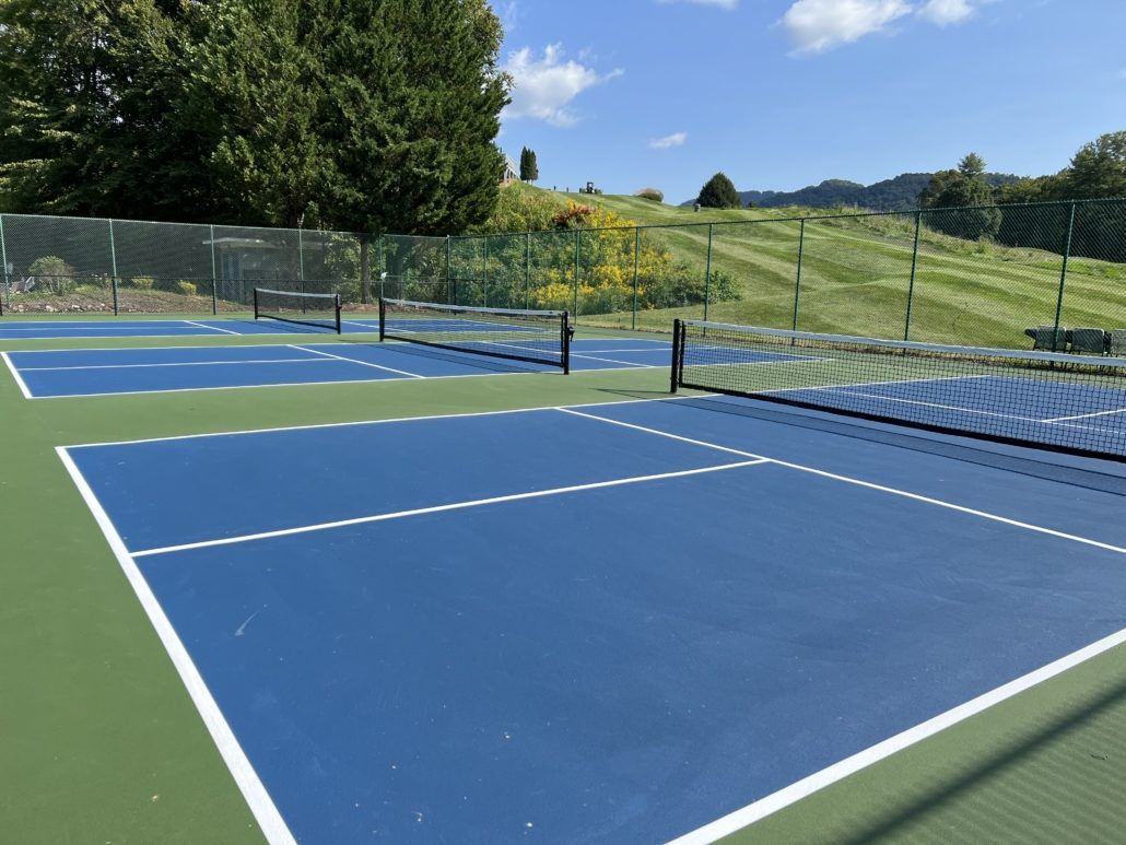 Blue pickleball courts with nets, green surrounds, and a hilly backdrop under a sunny sky.