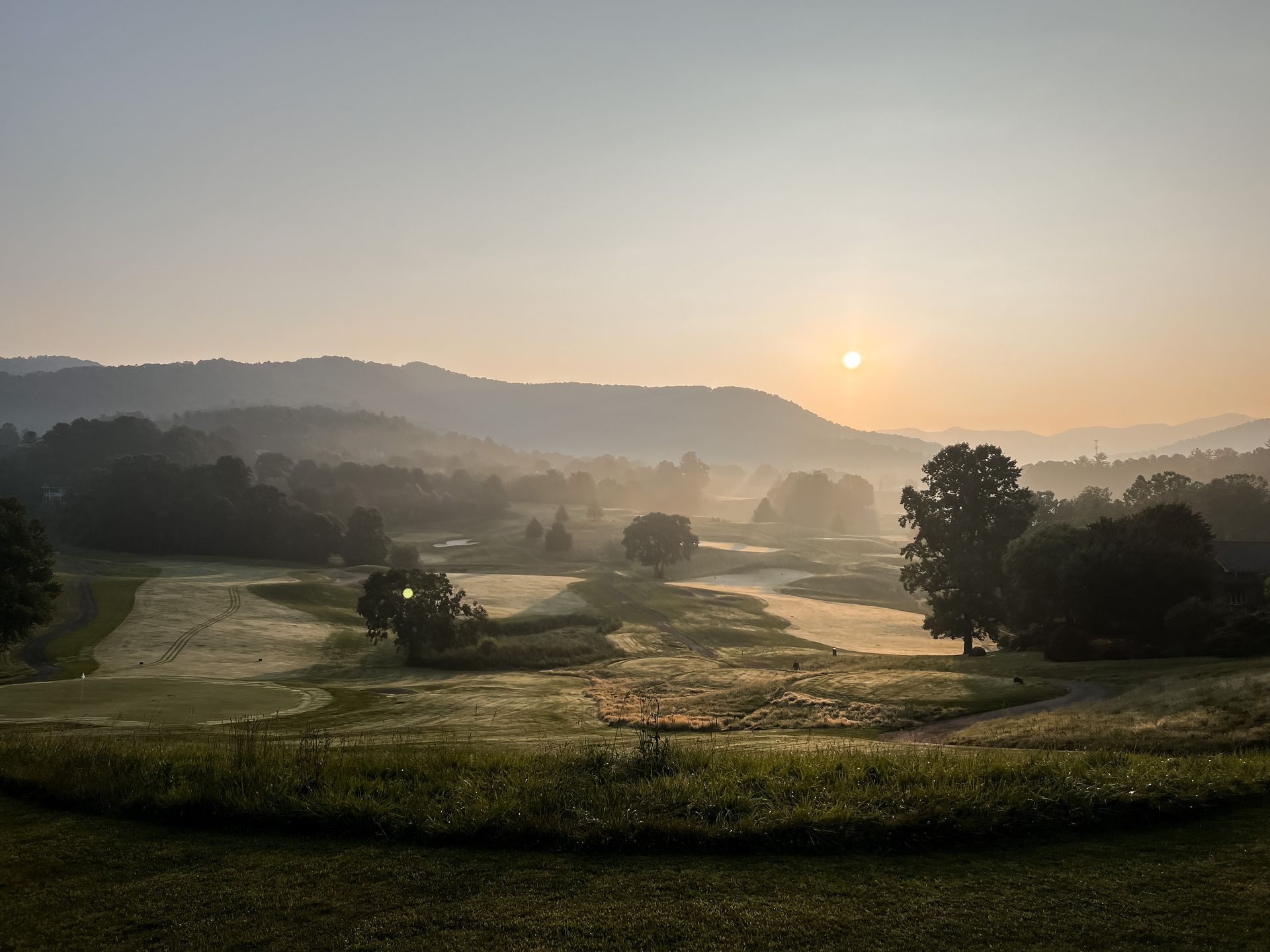 Sunrise over a misty valley with a golf course; trees dot the landscape; light-colored sky.
