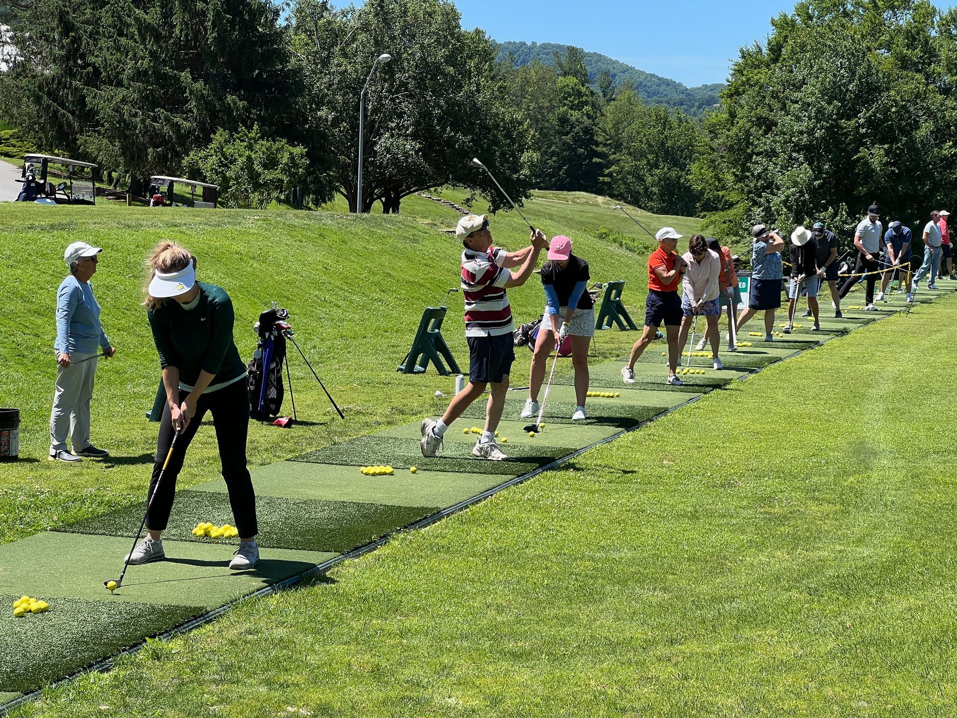 Golfers practicing swings on a driving range, sunny day. Green grass, blue sky, trees in the background.