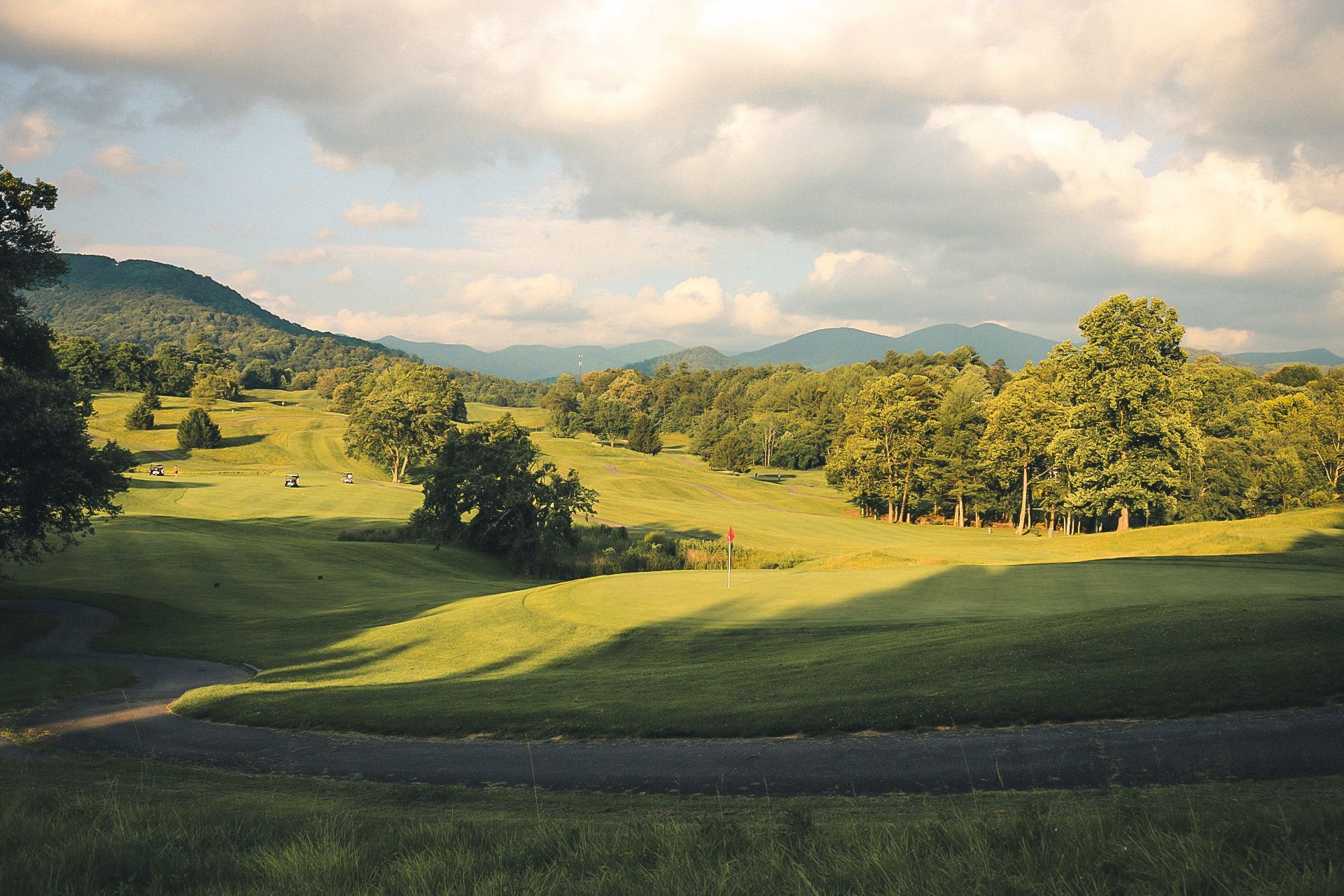 Green golf course with trees and distant mountains under a cloudy sky.