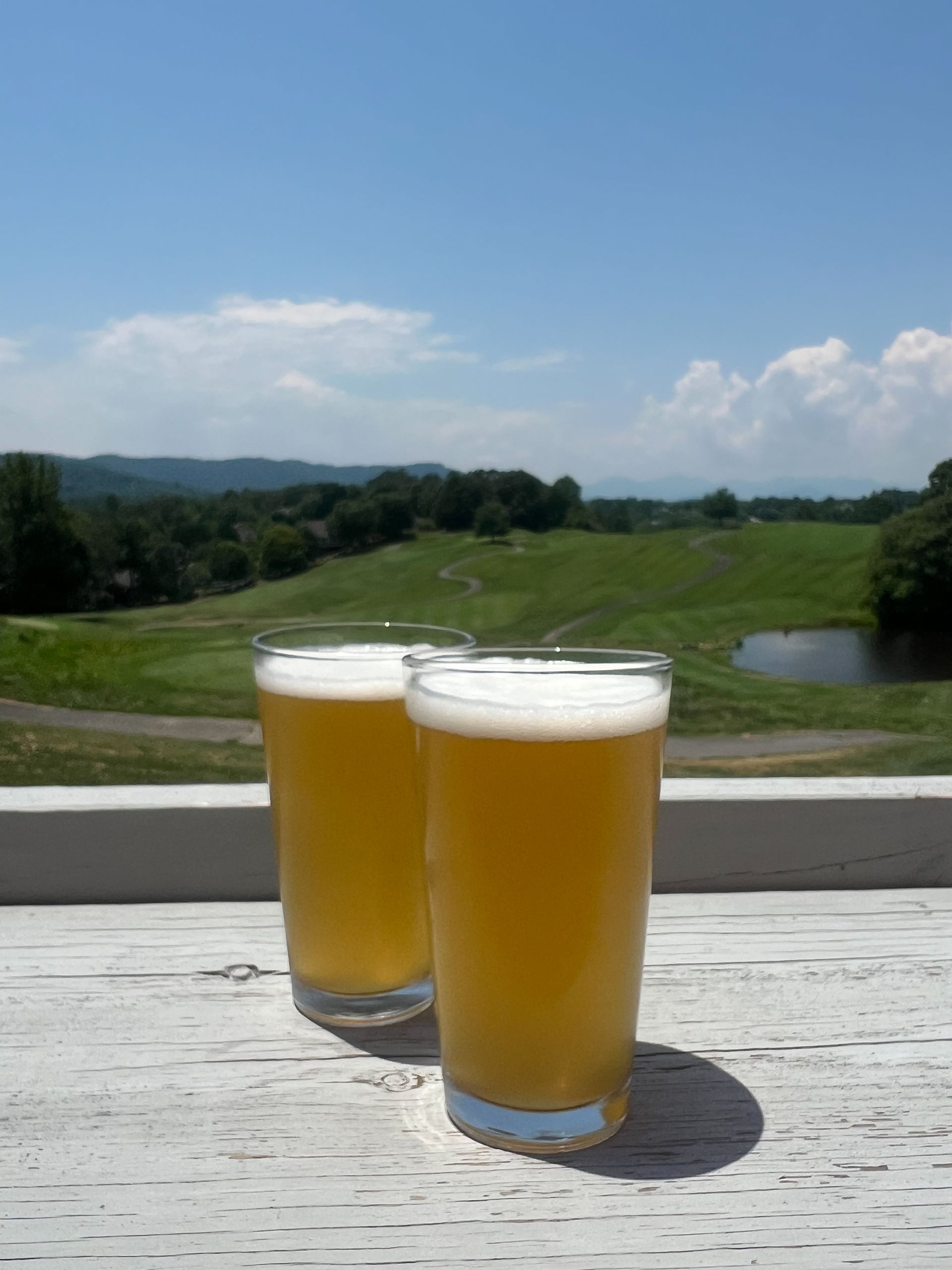 Two glasses of beer on a white surface, overlooking a green golf course and blue sky.