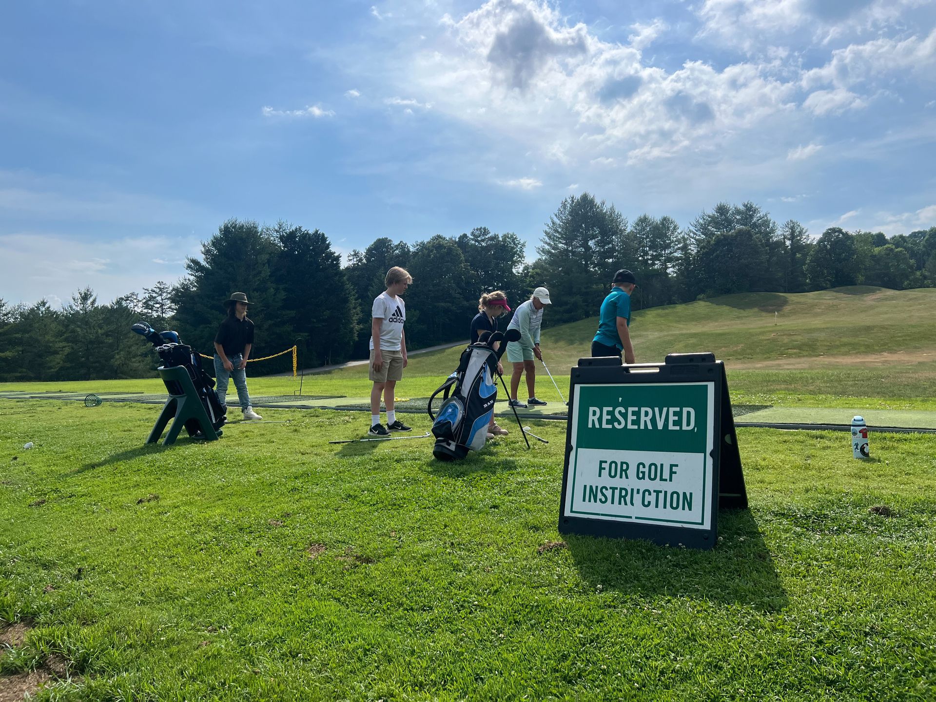 Golf lesson: People on a driving range with golf bags, reserved sign in front. Sunny day.