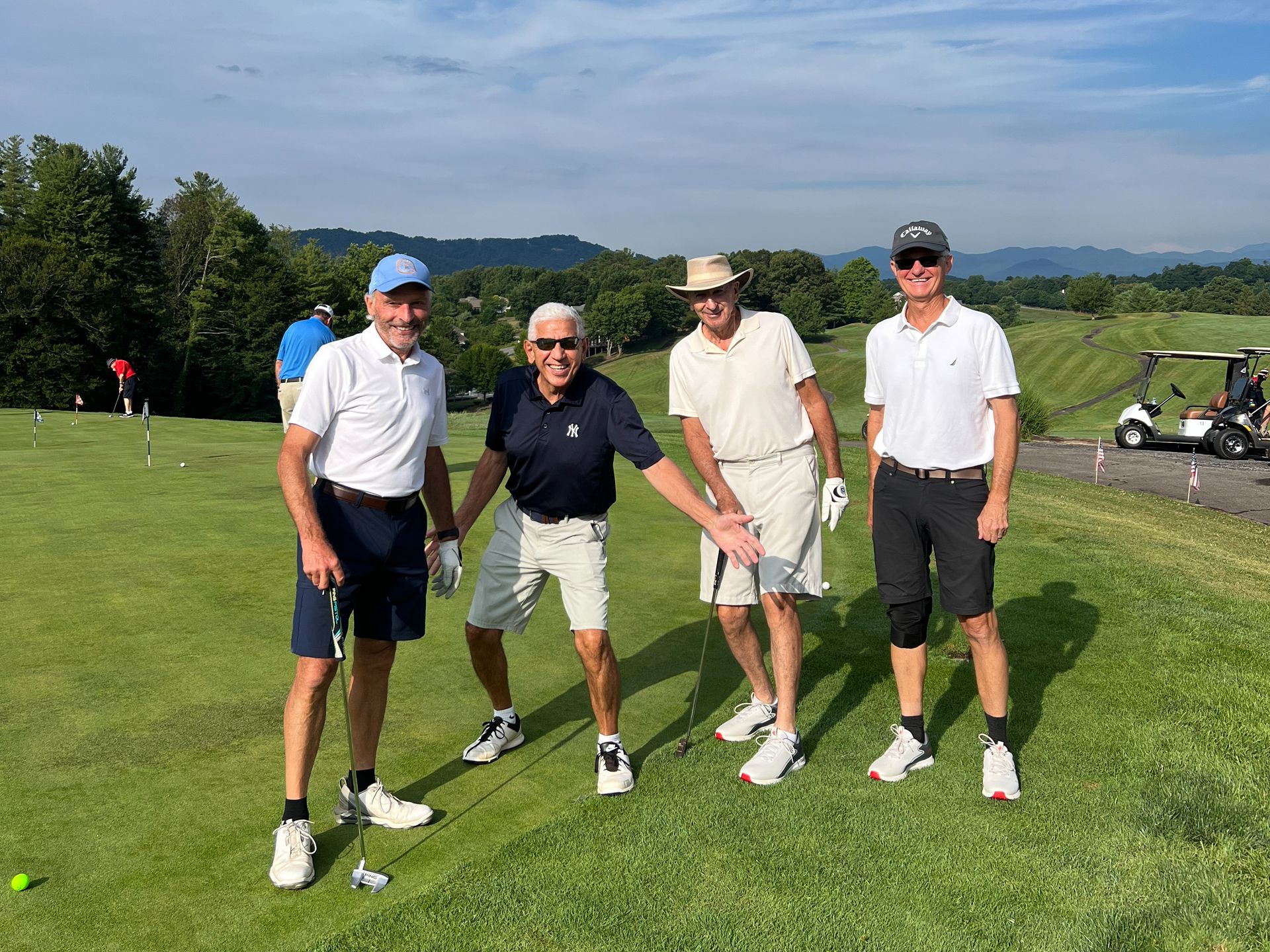 Four golfers on a green: one points, all smile. Golf course with hills and cart in background.