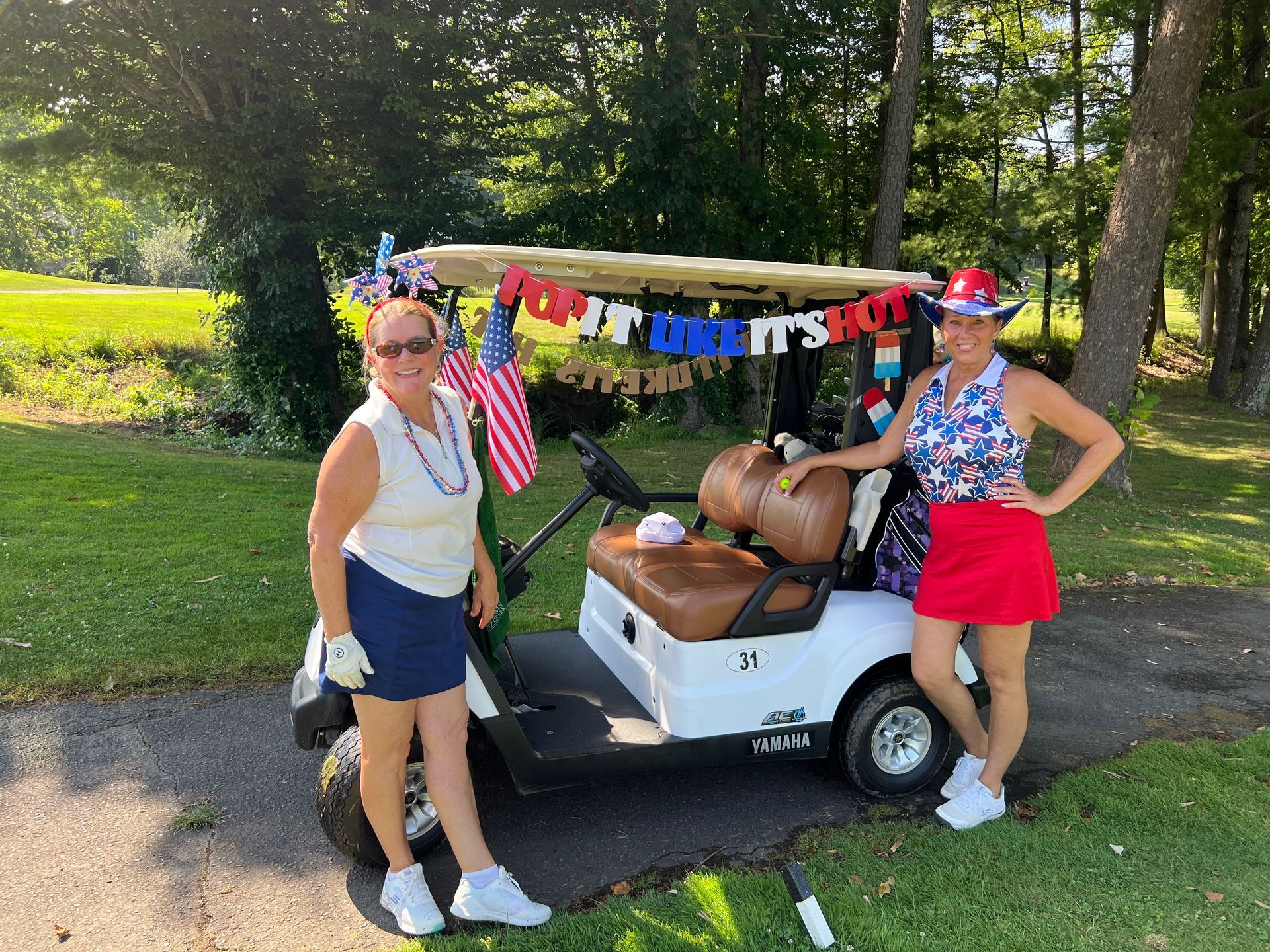 Two women pose with a golf cart decorated for a patriotic event in a sunny outdoor setting.