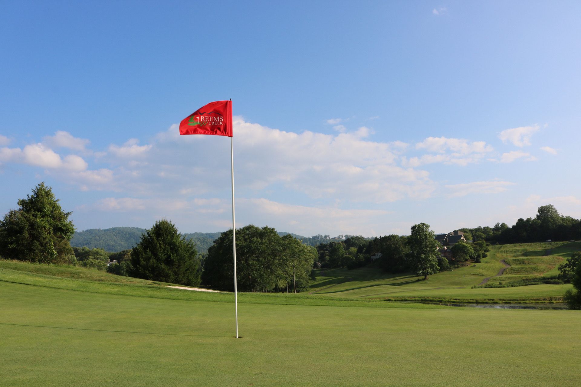 Red flag on a golf green under a blue sky, rolling hills in the background.
