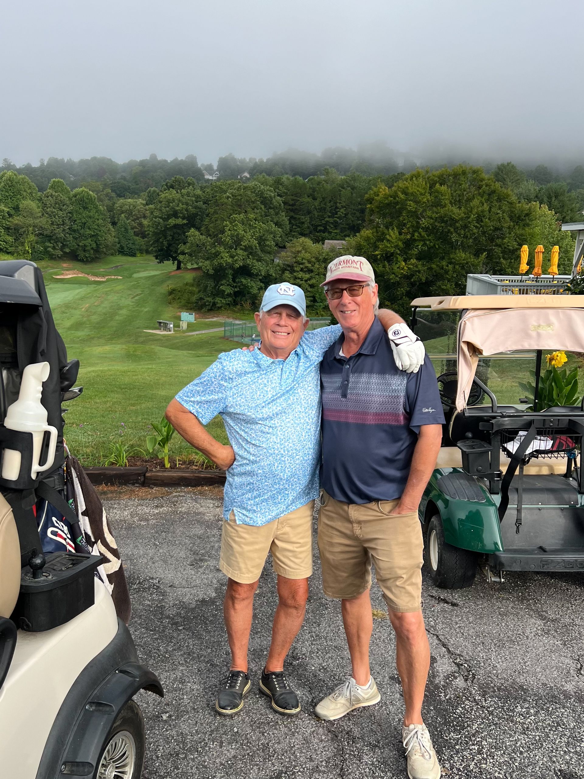 Two men in golf attire pose together, arms around each other near golf carts on a course.