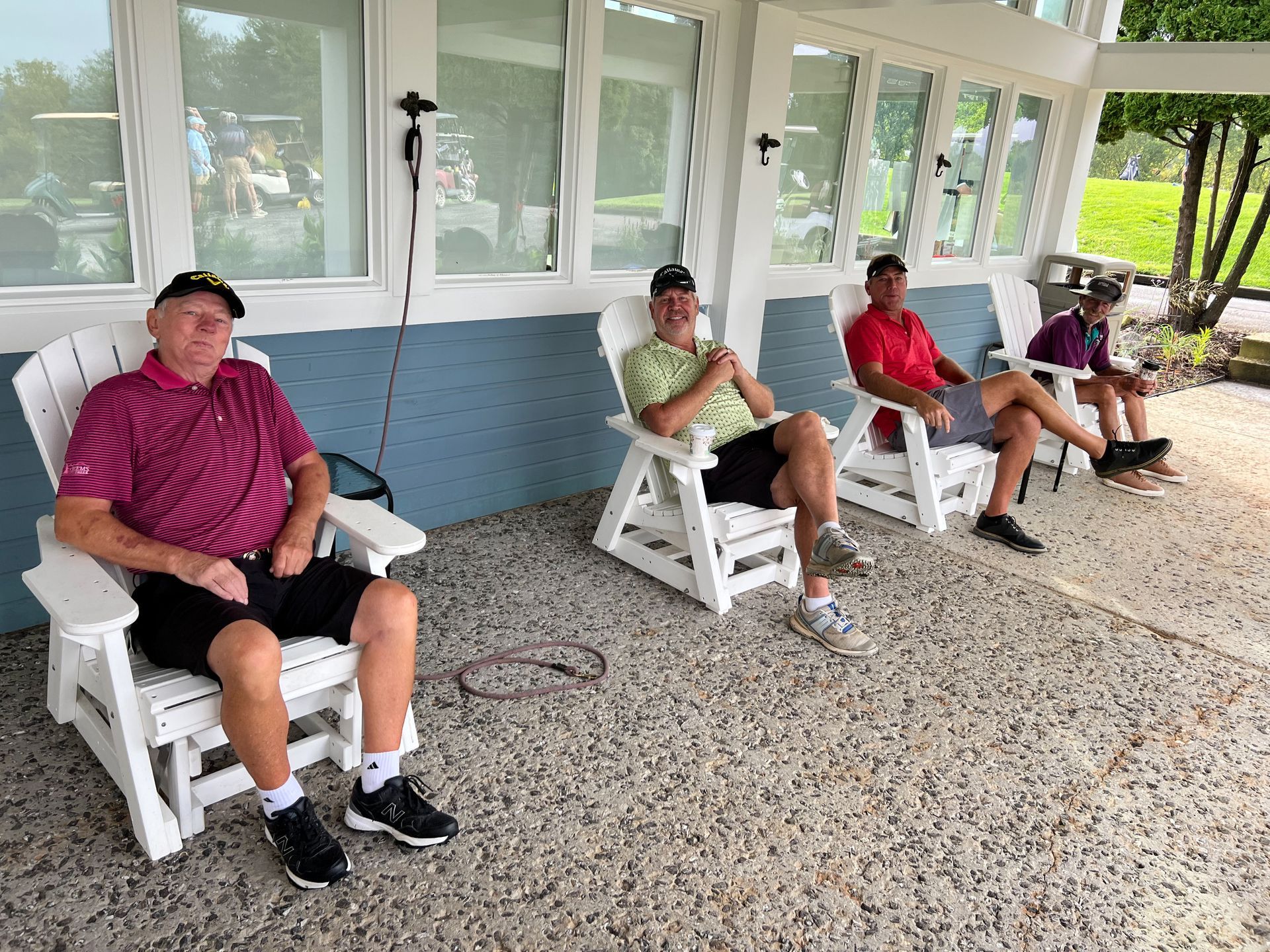 Four men in golf attire sit in white chairs on a patio.