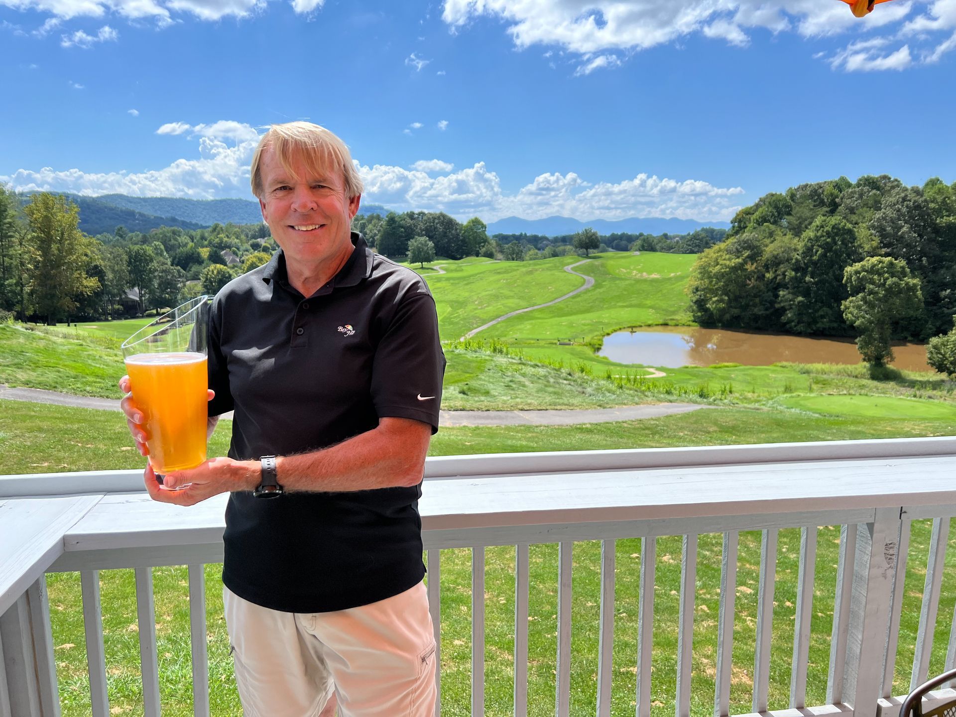 Man holding a beer on a deck overlooking a green field and mountains under a blue sky.