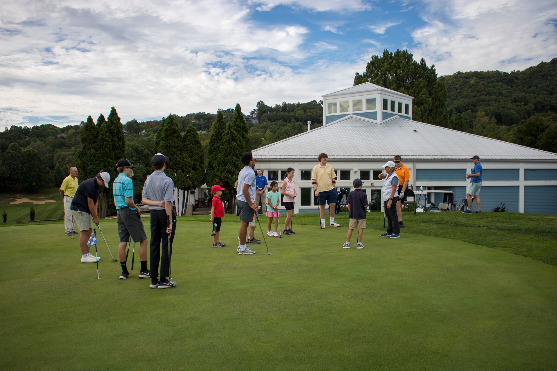 People putting golf balls on a green, near a clubhouse under a cloudy sky.