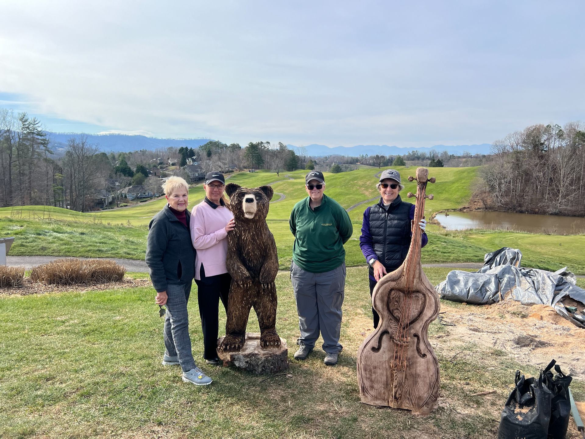 Four people stand near a bear and bass wood carvings outdoors, mountains in the background.