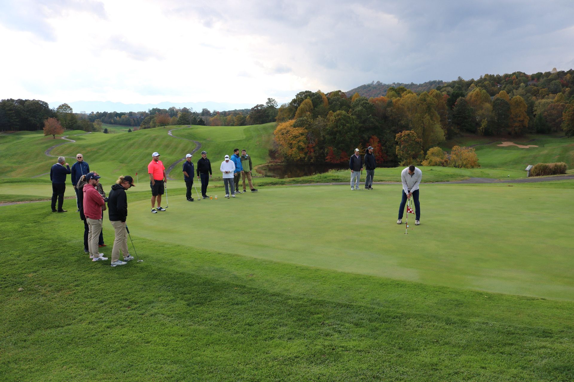 A golfer putts on a green, watched by a group of people on a golf course. Overcast day.
