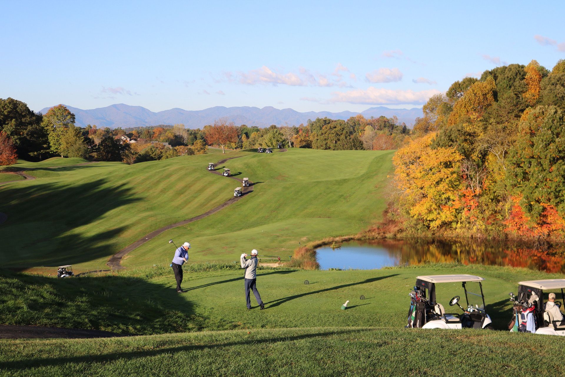 Golfers teeing off on a fairway; fall foliage, pond, and golf carts visible. Mountains in the distance.
