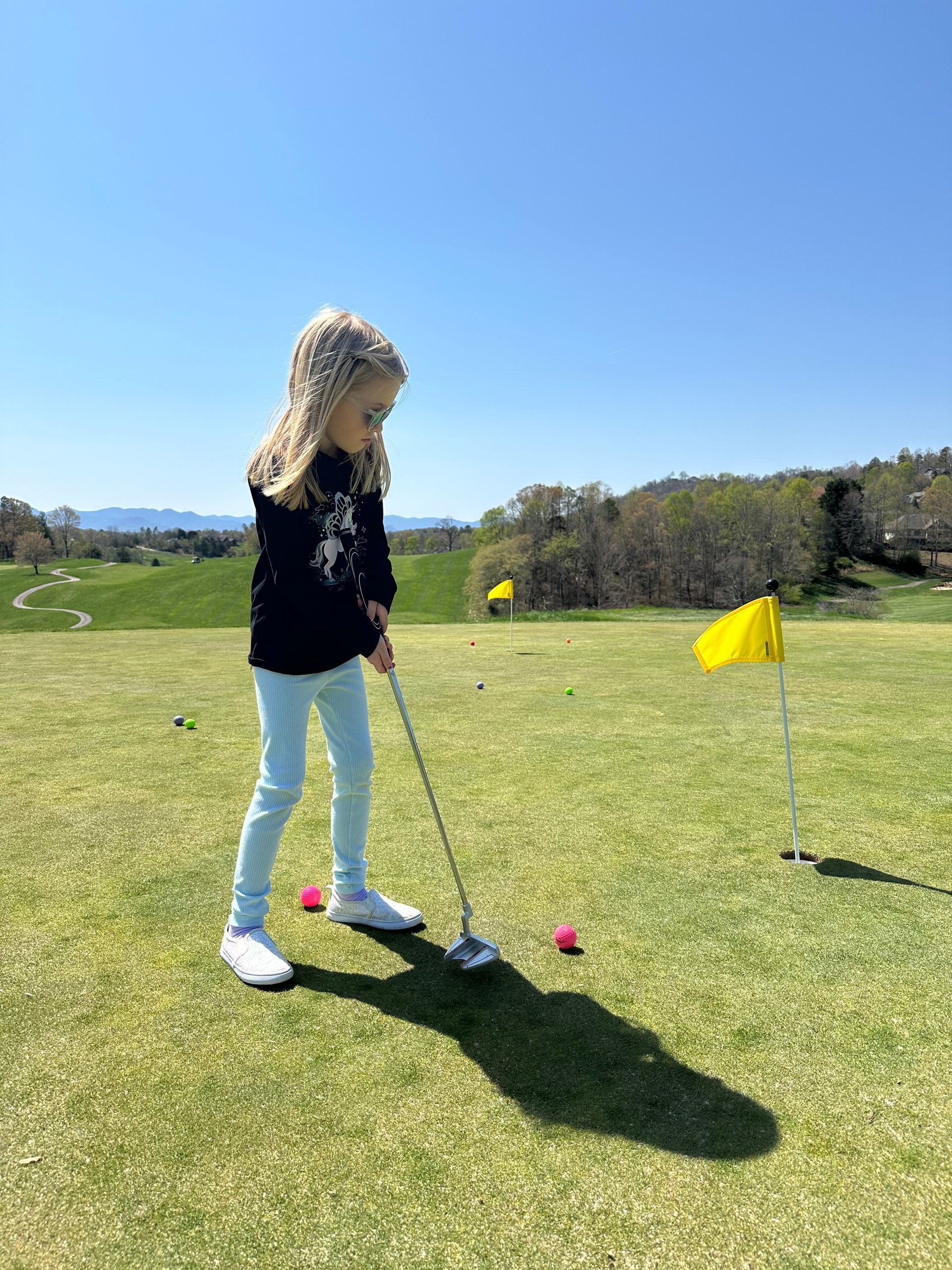 Young girl playing golf on a sunny green, aiming for the hole with pink golf balls nearby.