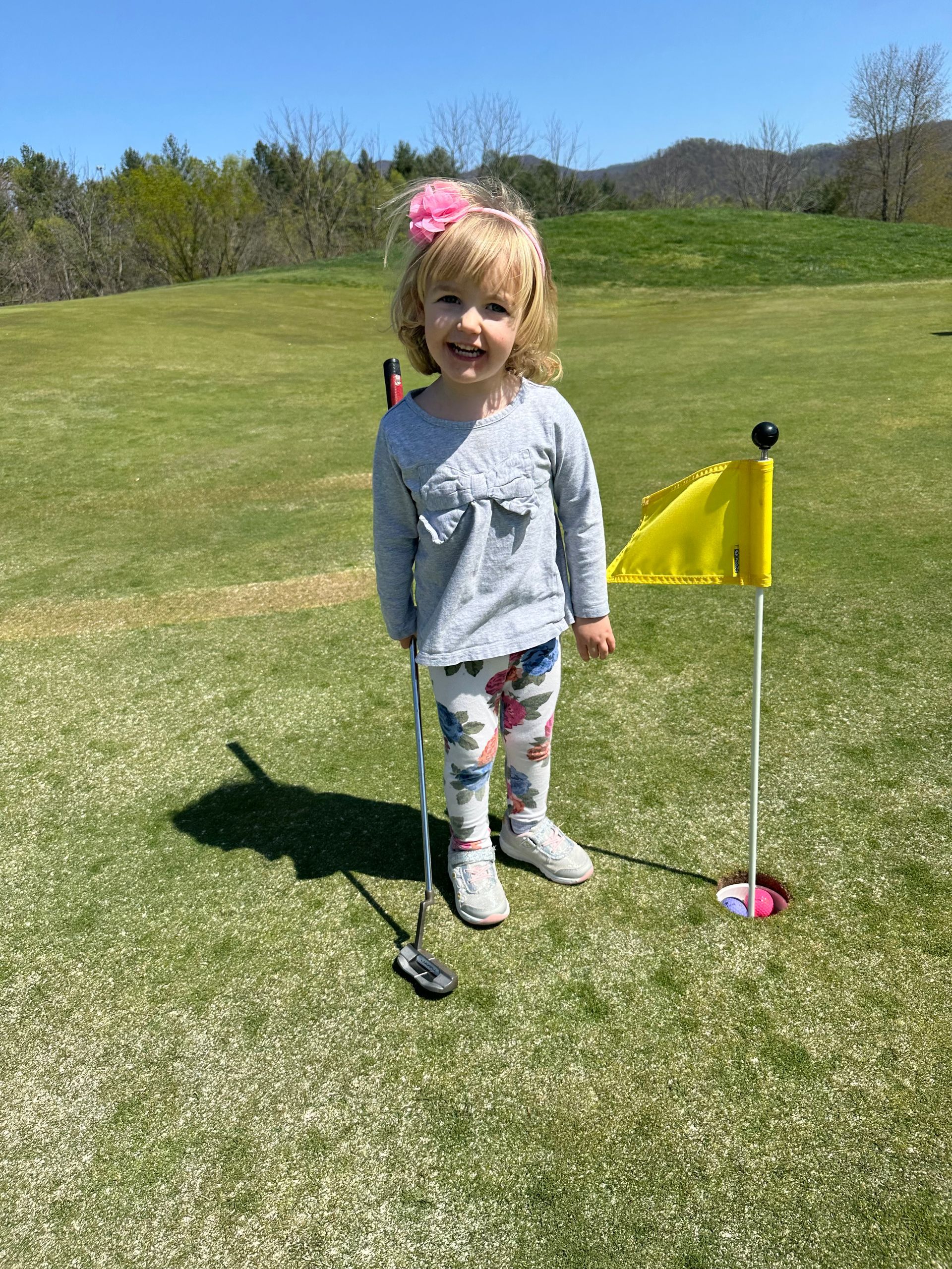 Young child smiles on a putting green, holding a golf club near a yellow flag and hole.