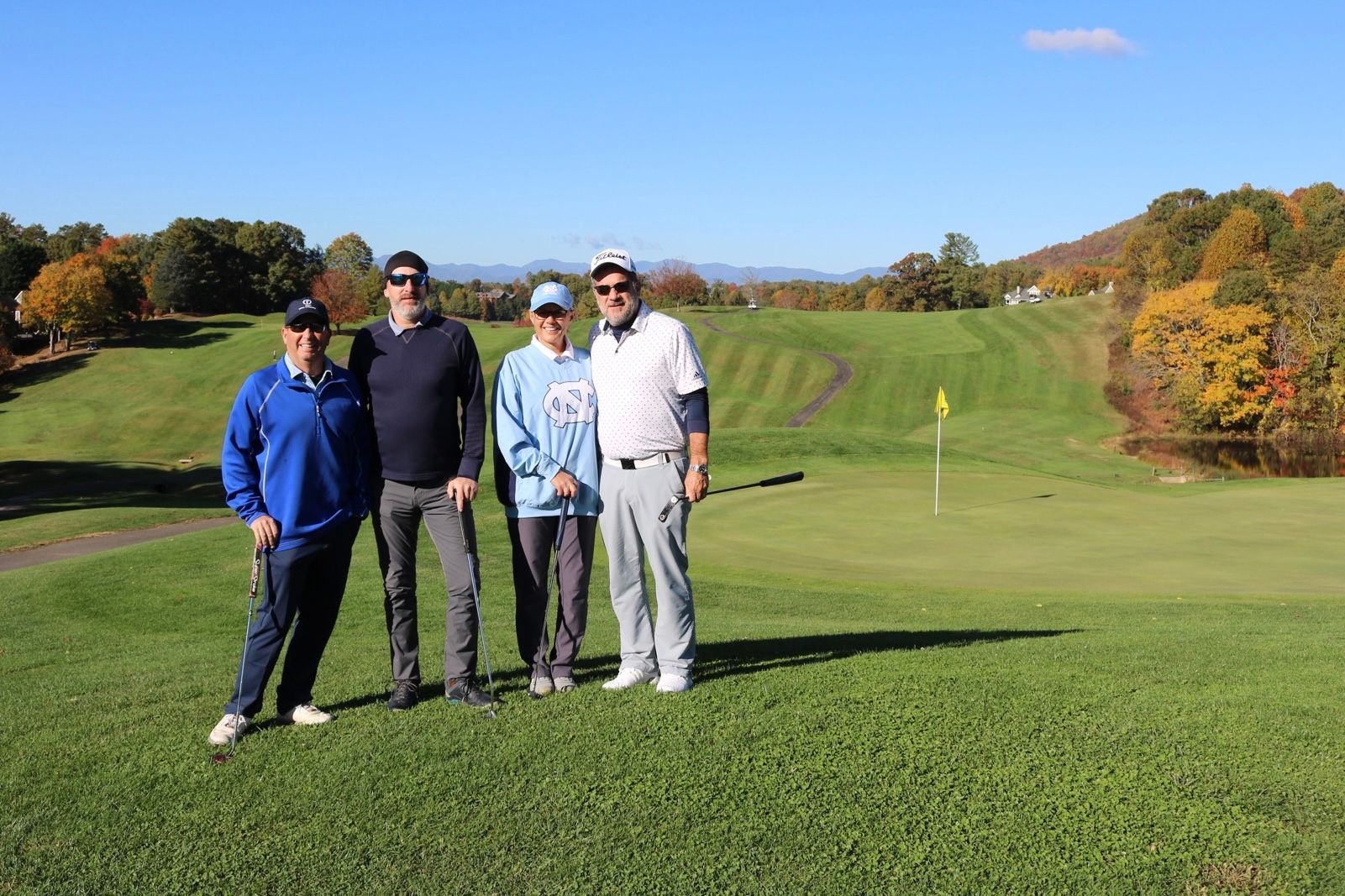 Four people pose on a golf course, green grass, and mountains in the distance.