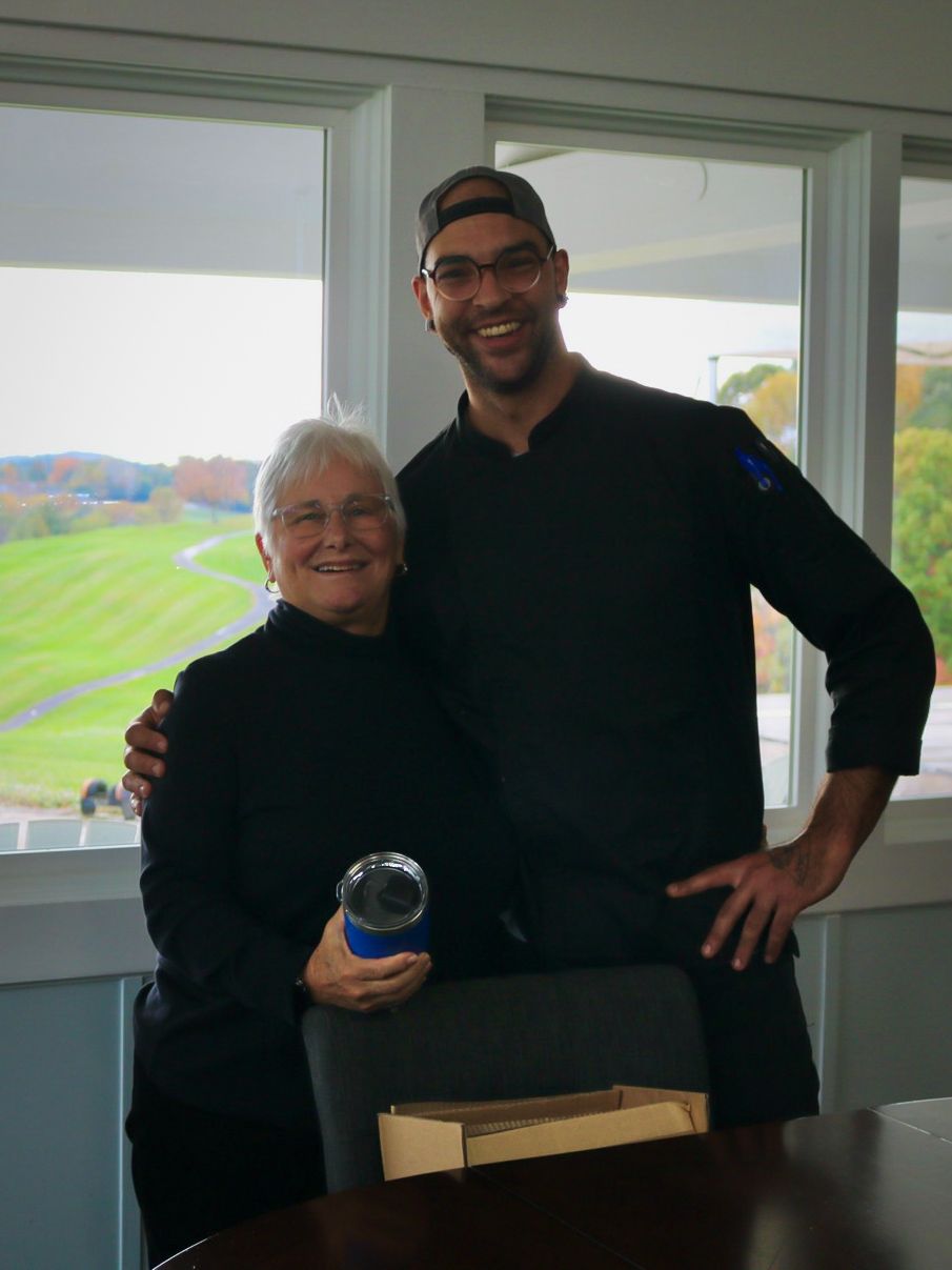 An older woman and a man in a chef's coat pose together, smiling, near a window overlooking a green landscape.