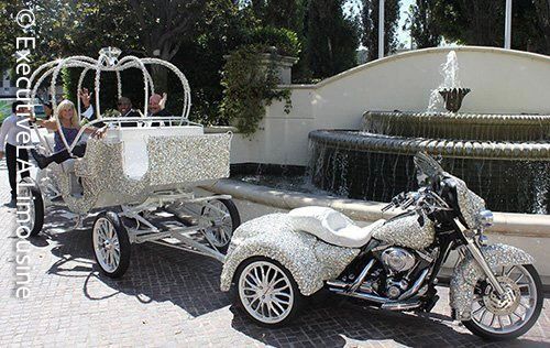 White motorcycle towing a small silver carriage with heart-shaped arches beside a fountain outdoors